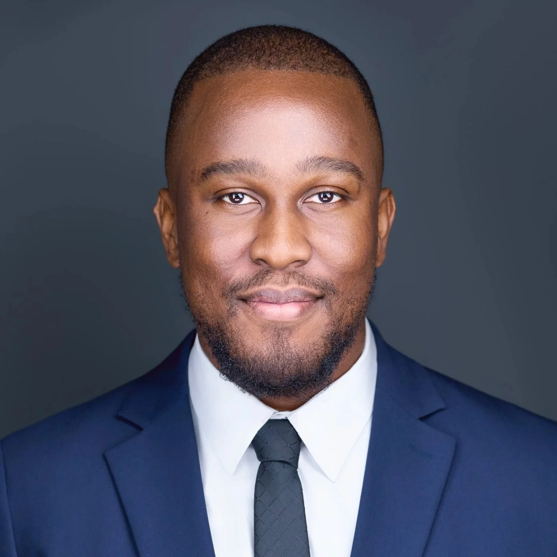 Business headshot of business executive in a dark blue suit, white shirt, and black tie, smiling against a dark grey background.  Jessica Mills Photography
