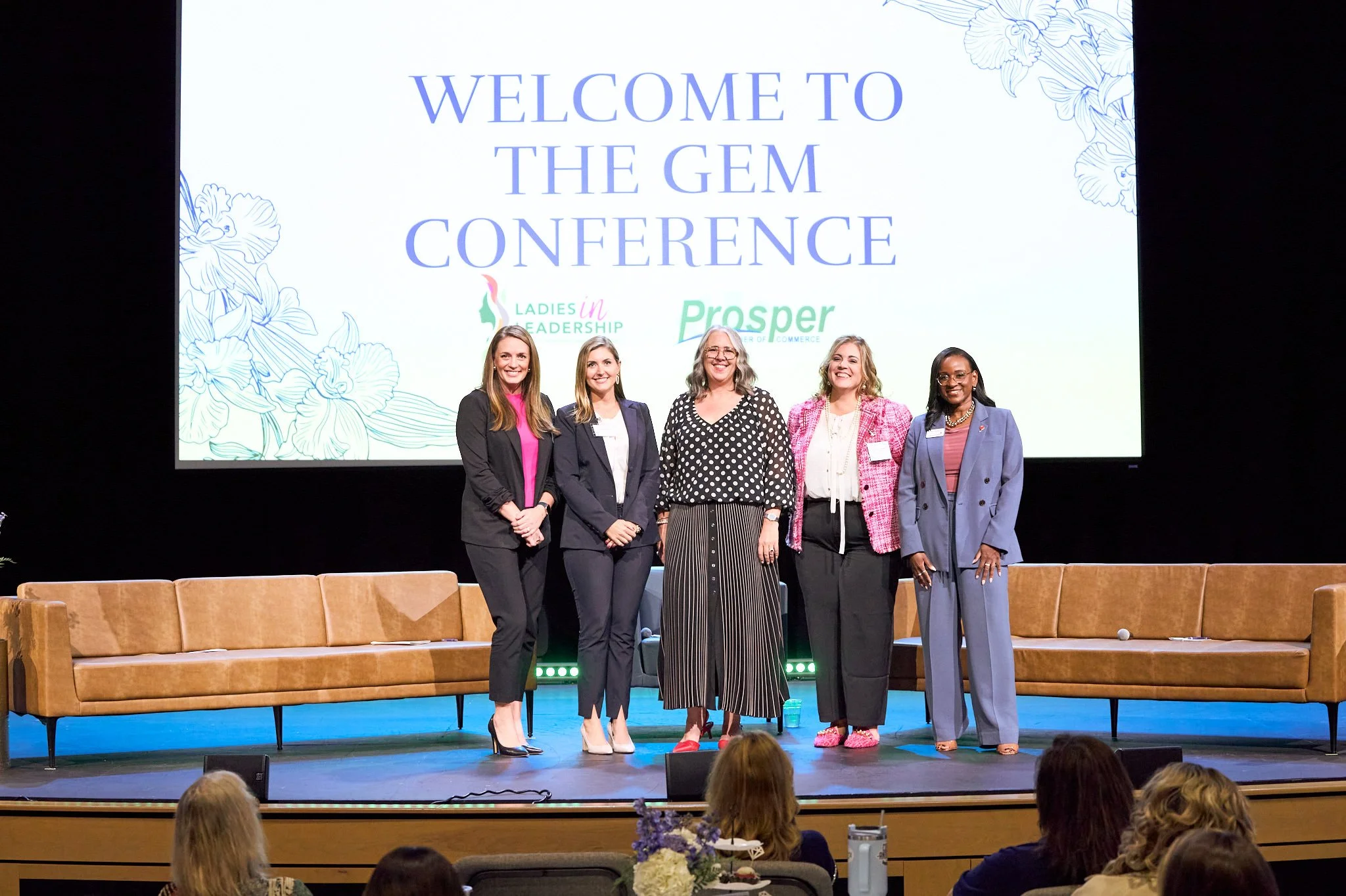 Group of five women standing on stage in front of a large screen that reads 'Welcome to the GEM Conference. Ladies in Leadership, Prosper,' with floral illustrations, during a professional event.