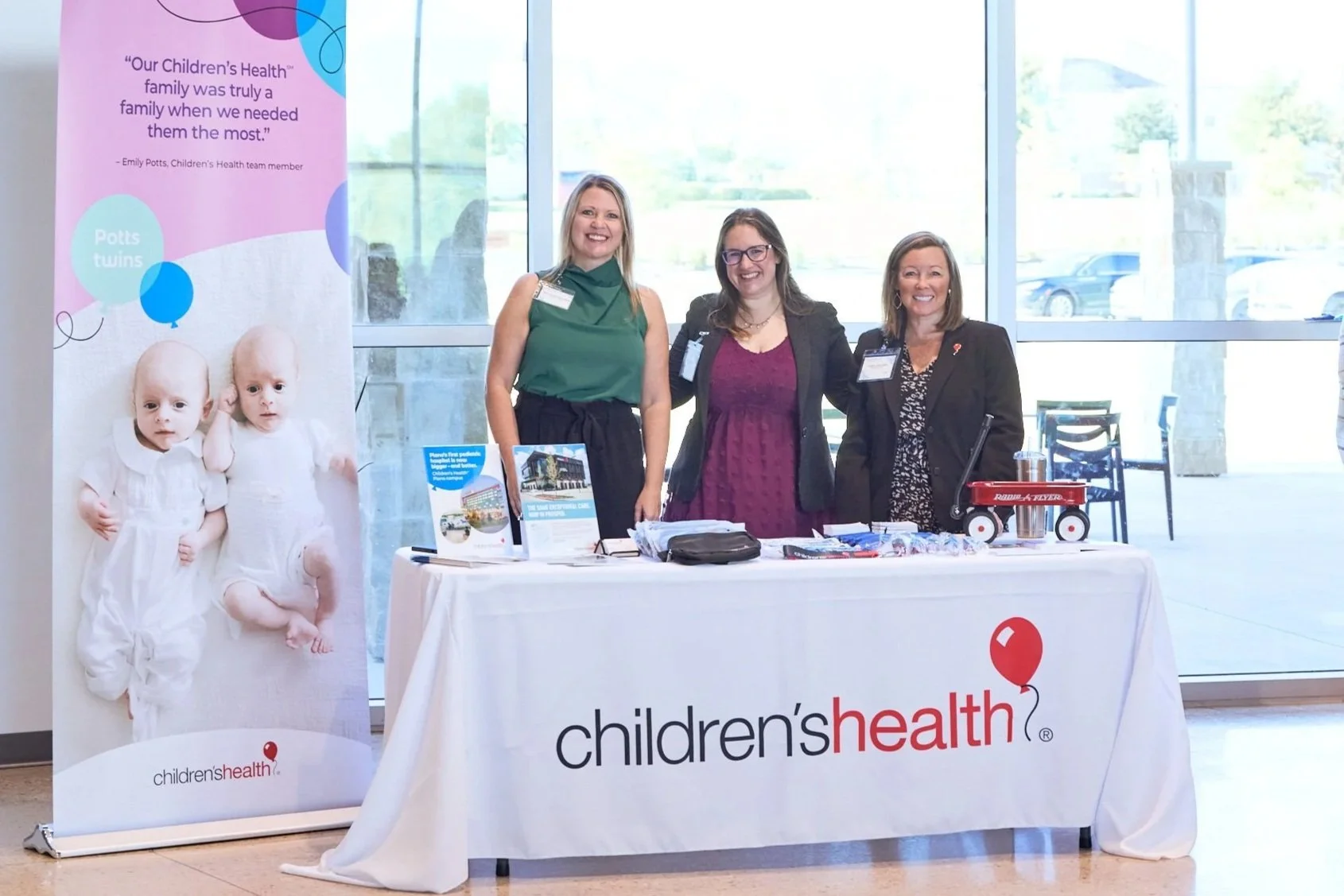 Three women standing behind a table with children's health materials at an event, with a children's health banner featuring twins and quotes about family health.