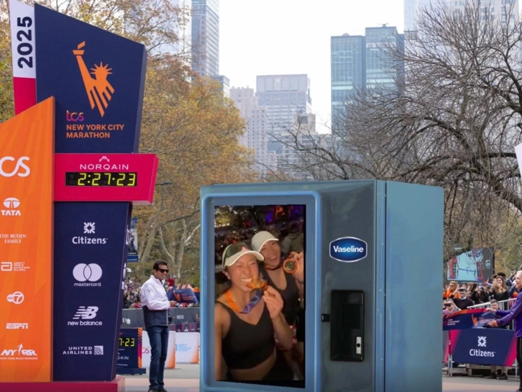 Two women taking a selfie inside a phone booth near the New York City Marathon finish line, with race banners, onlookers, and city buildings in the background.