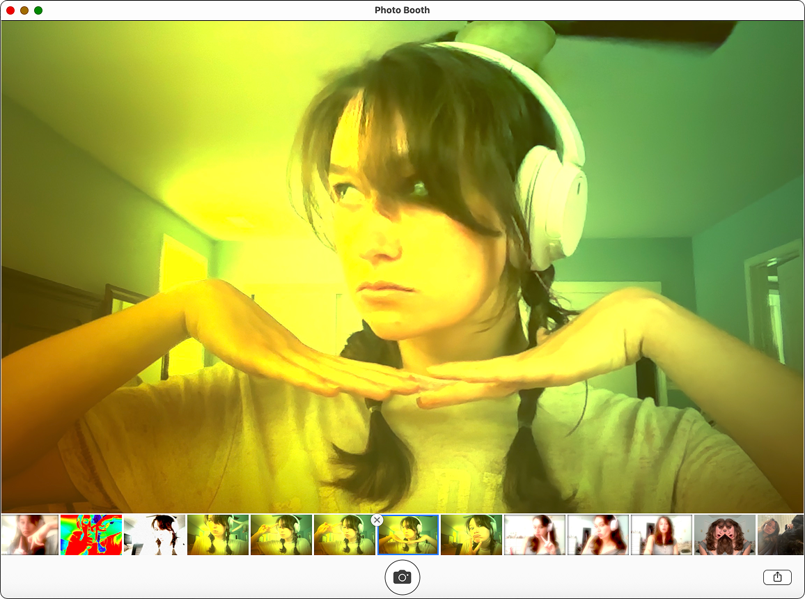 Young woman with dark hair, wearing headphones, making a playful pose with her hands under her chin, in a room with green and yellow lighting.