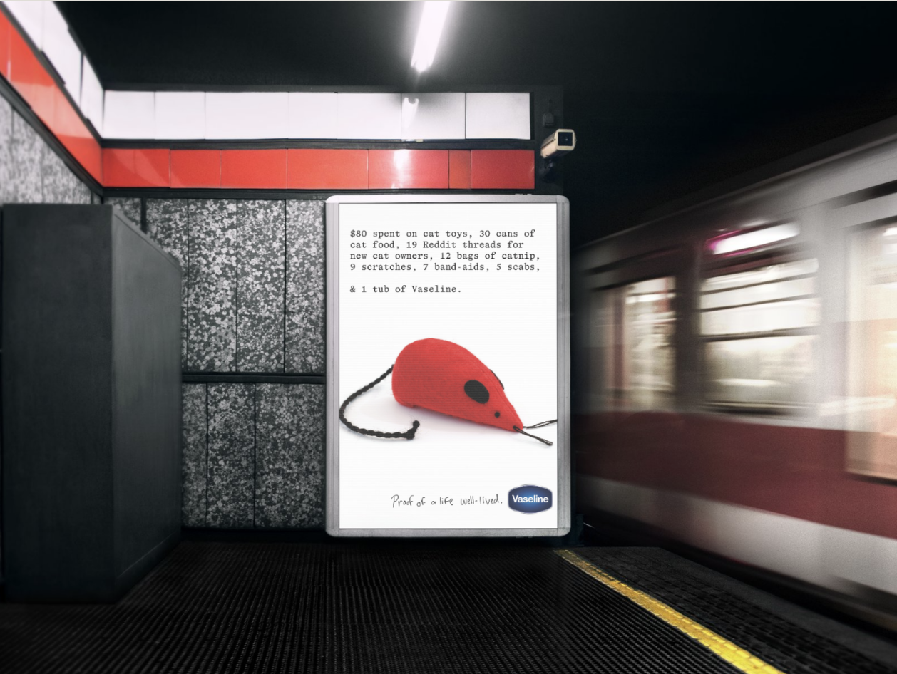 Digital billboard on subway platform displaying a red toy mouse with a black tail and eye, with text about purchases made for a cat, including cat toys, food, and supplies, and a caption 'Proof of a life well-lived.'