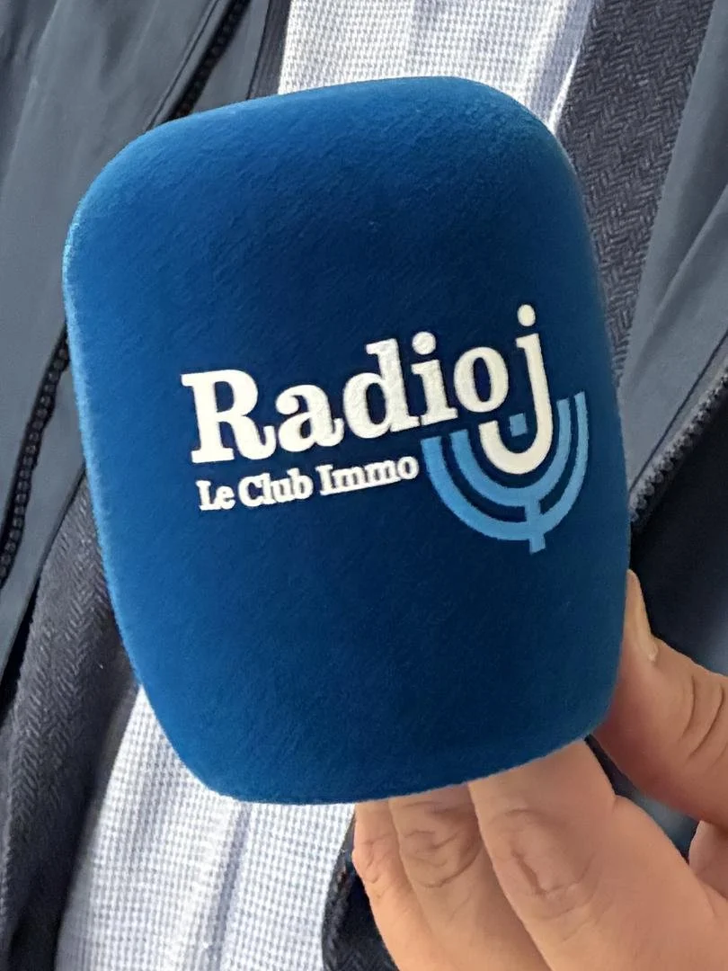 Julie Douarin et Marc Ezrati, assises à une table de radio, avec des microphones et des écouteurs devant eux, dans un studio radiophonique Radio J, avec des écrans derrière affichant le logo Radio J et la fréquence 94,8 FM.