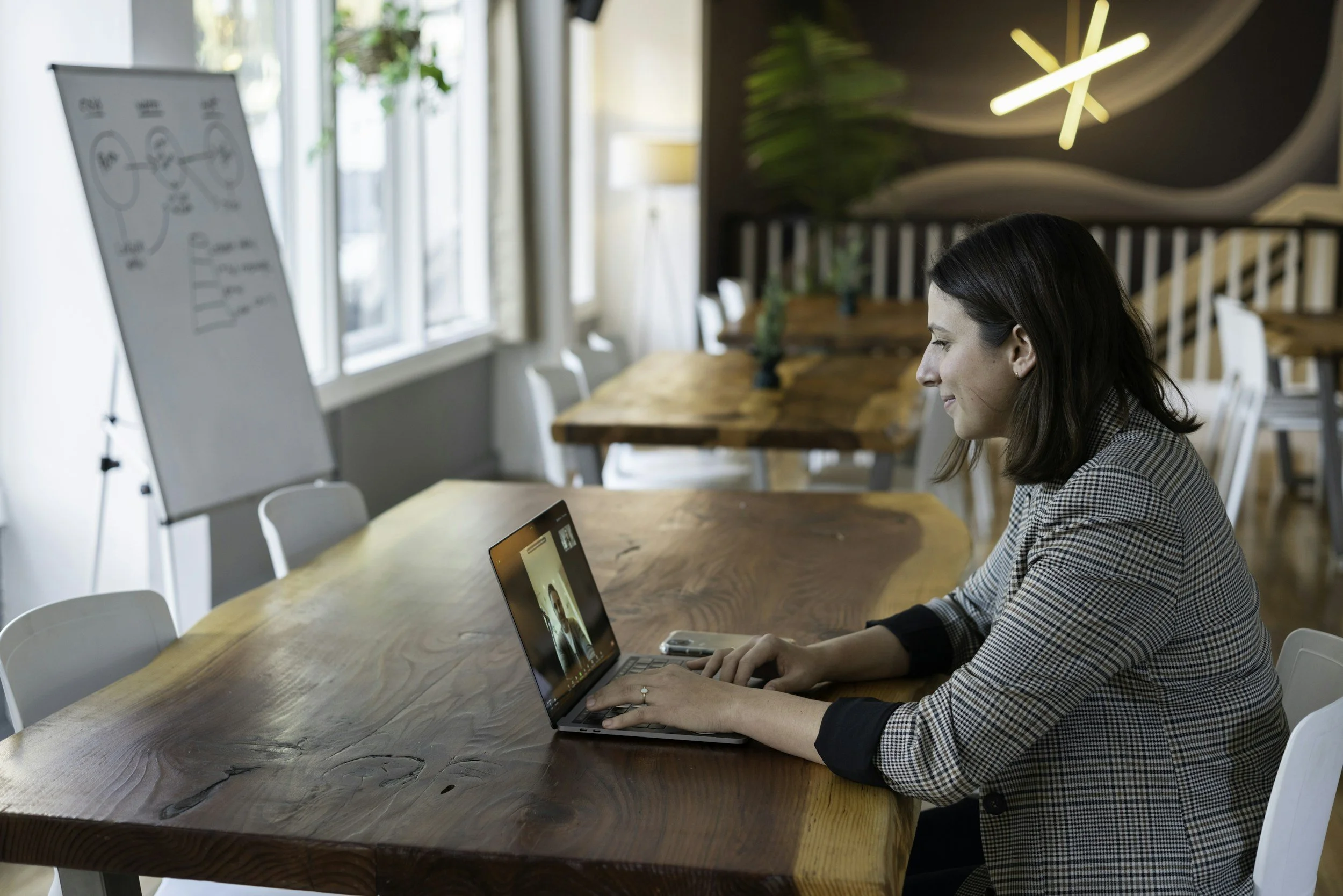 Woman in a checkered blazer video conferencing on a laptop at a wooden table in a modern office with natural lighting and a whiteboard in the background.