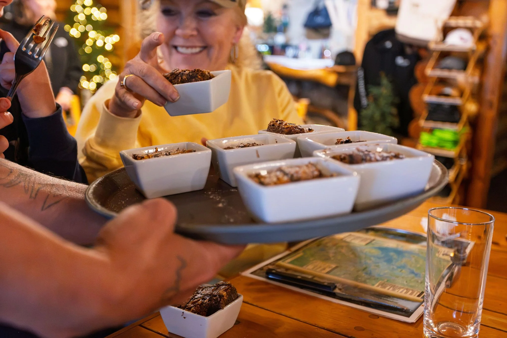 A woman in a yellow top smiling and holding a small white bowl of dessert, with several similar bowls on a tray in front of her. She is in a cozy, decorated interior with warm lighting and blurred background, possibly a restaurant or café.
