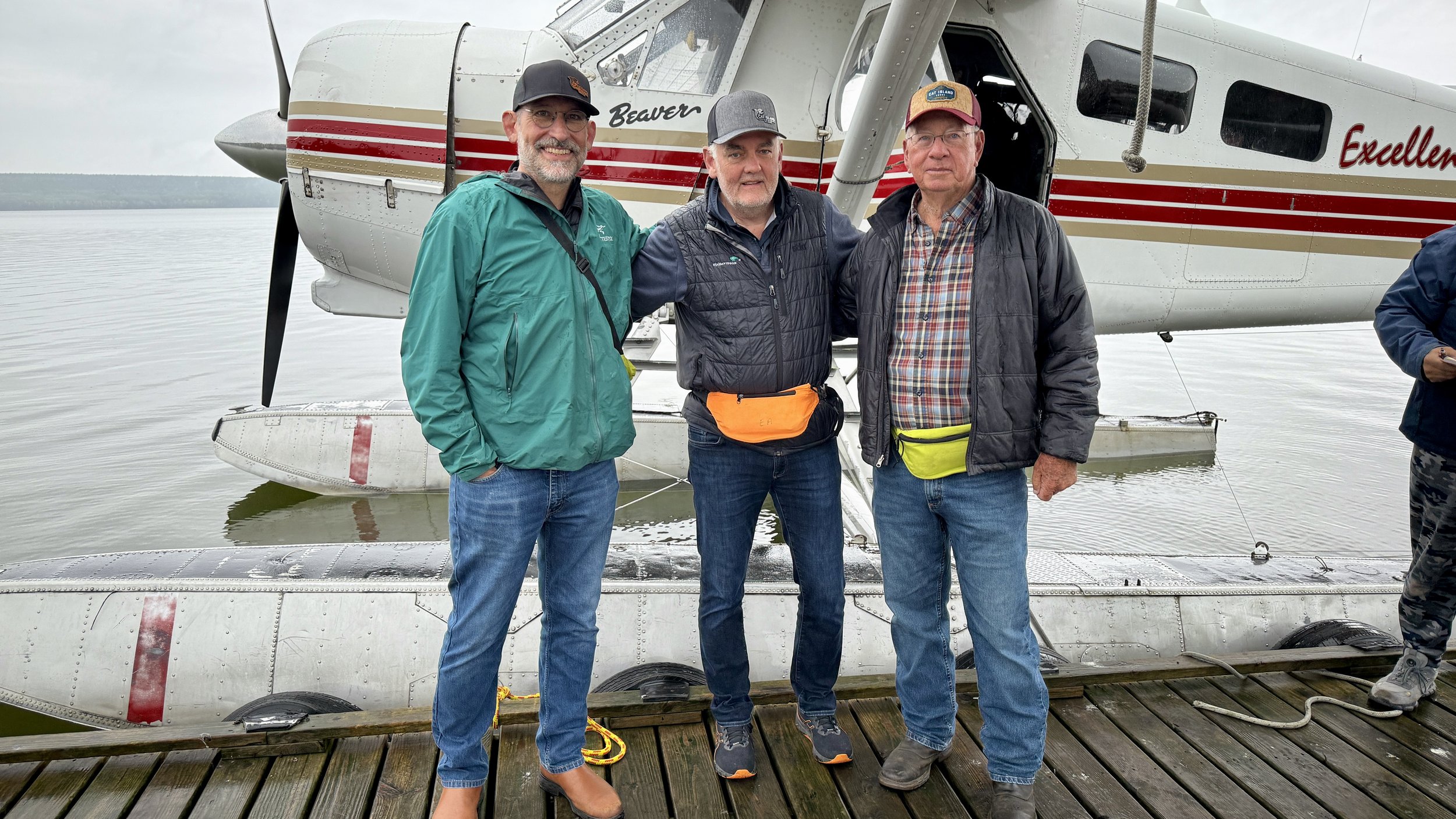 Three men standing on a wooden dock at Cat Island Lodge in front of a white float plane with red and gold stripes, with water and a cloudy sky in the background.