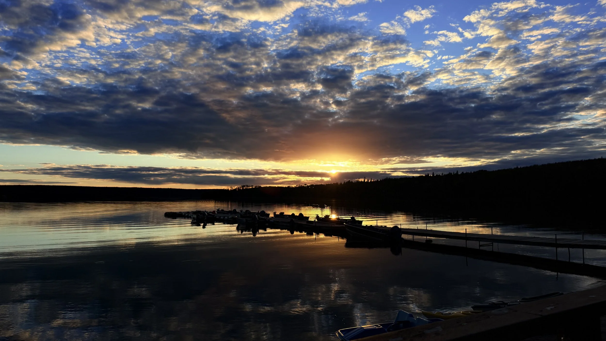 Sunset over a calm lake with a partly cloudy sky, silhouetted trees in the distance, and docks with boats along the shoreline.