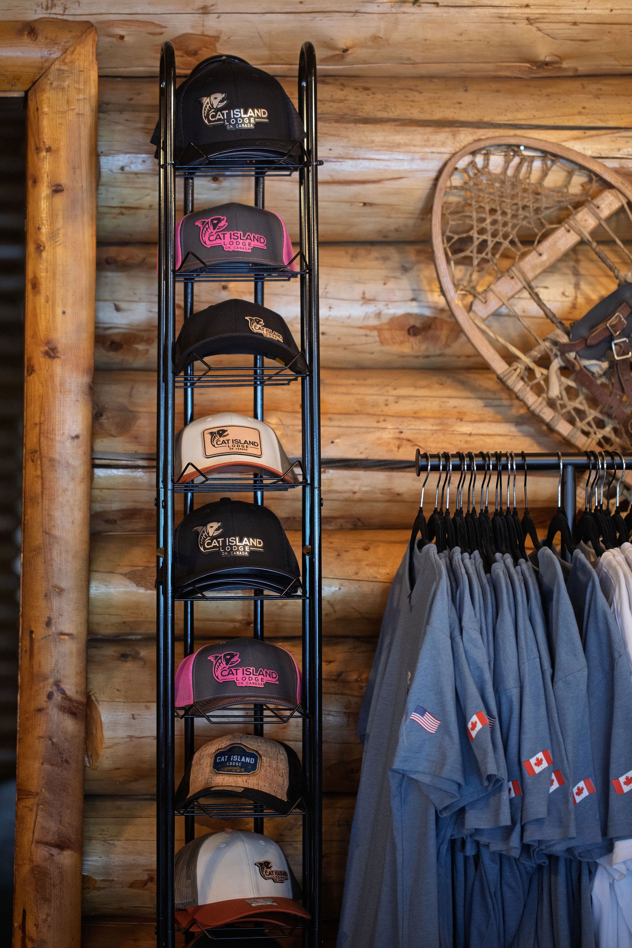 A display rack with multiple baseball caps featuring the 'Cat Island Lodge' logo. The caps are in various colors, including black, pink, and beige. To the right, a rack of gray shirts with small Canadian flags and red maple leaves is visible. The bac