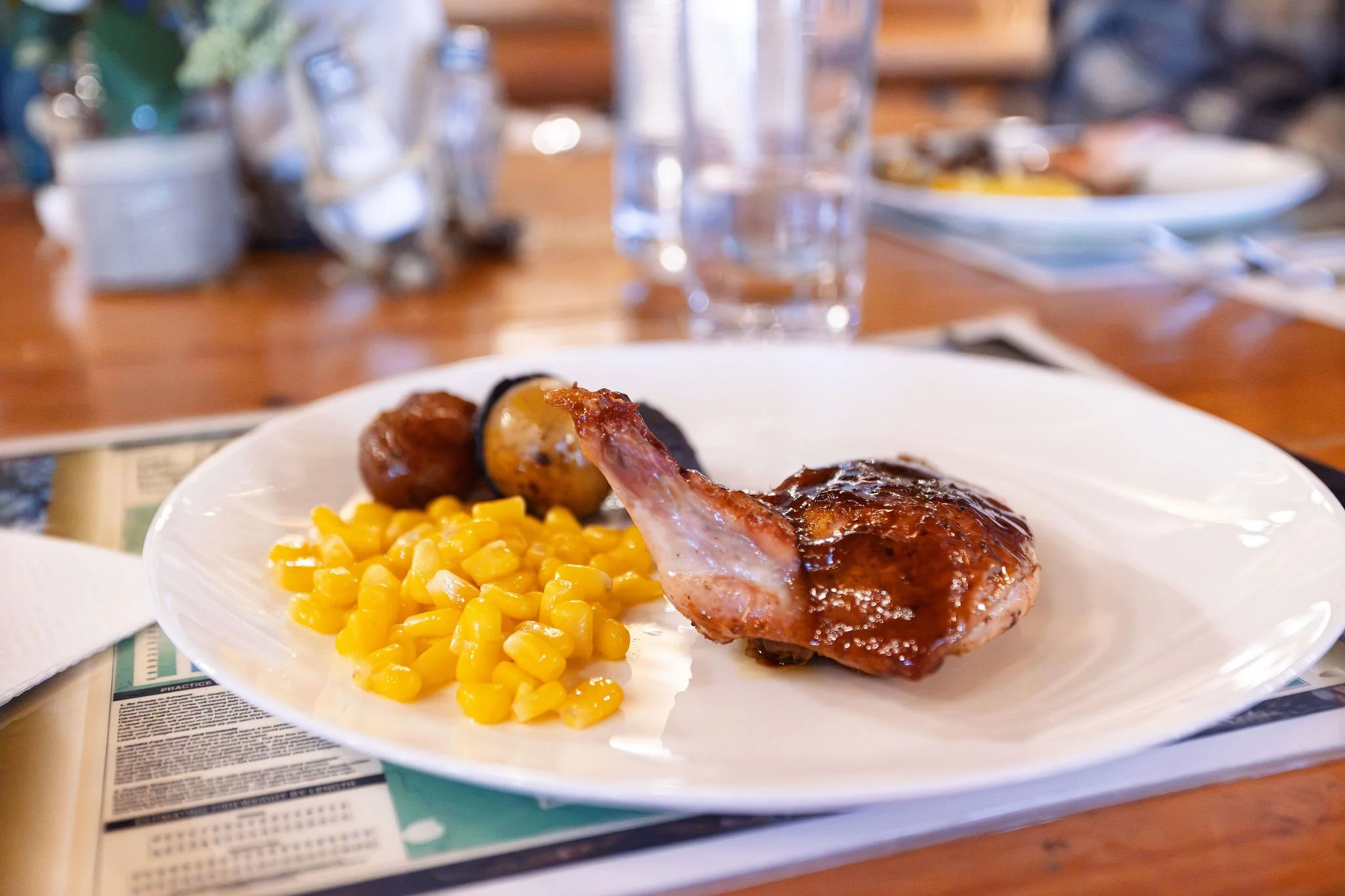 A white plate with a serving of corn, two meatballs, and a piece of glazed meat, possibly pork or chicken, with a brown sauce, on a table with glasses and utensils in the background.