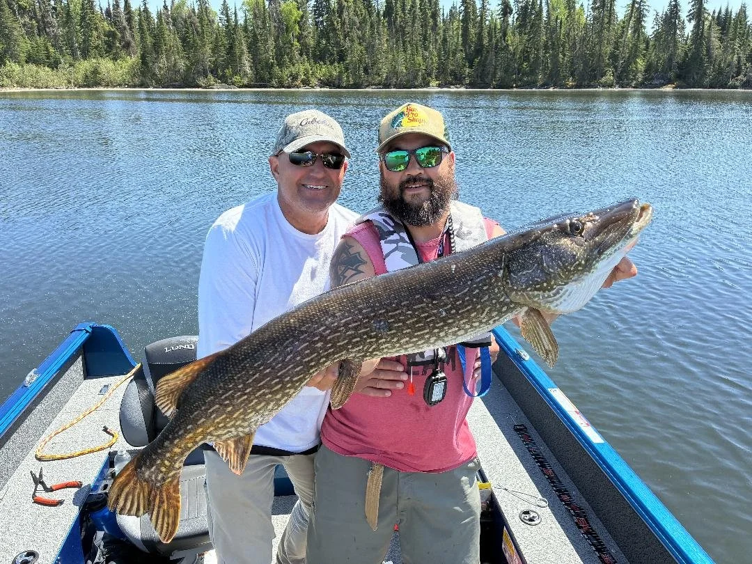 Two men on a boat holding a large fish in front of a lake with a forest in the background.