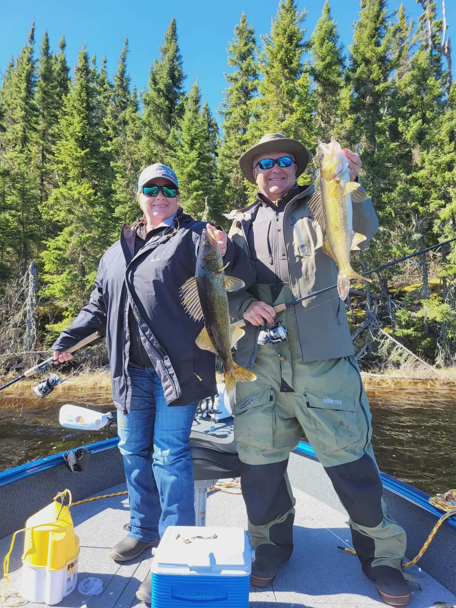 Two people on a boat holding caught fish with a forest background and clear blue sky.