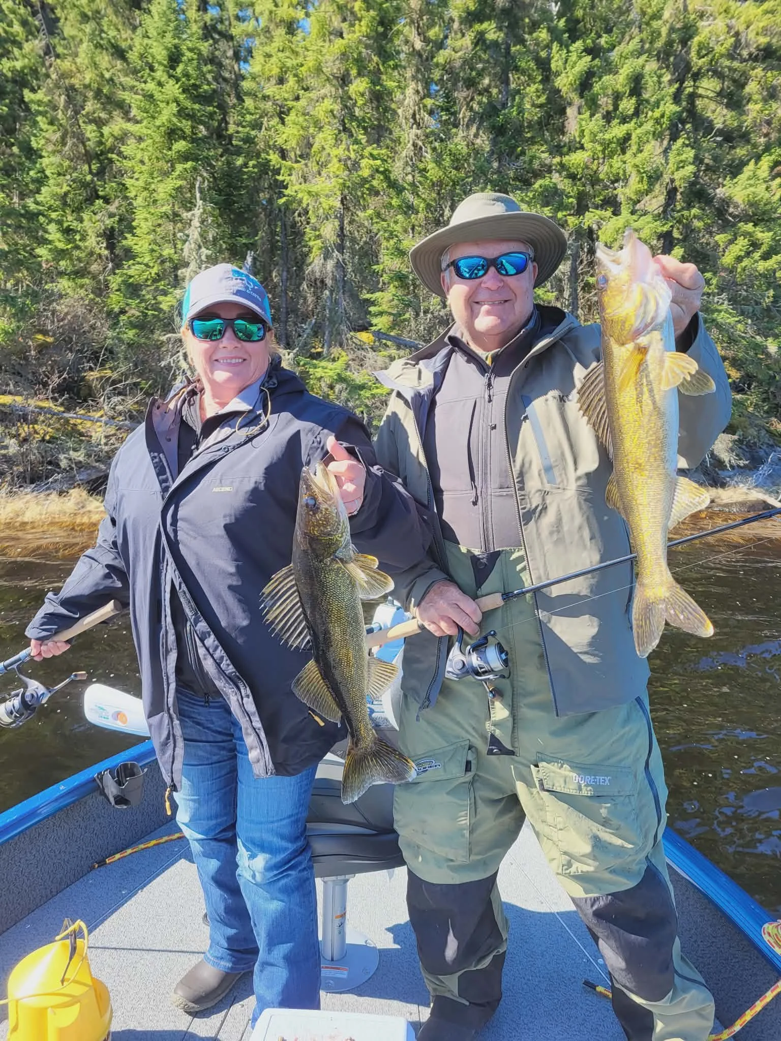 Two people on a boat holding up large fish they caught, with a backdrop of lush green trees and a clear sky.