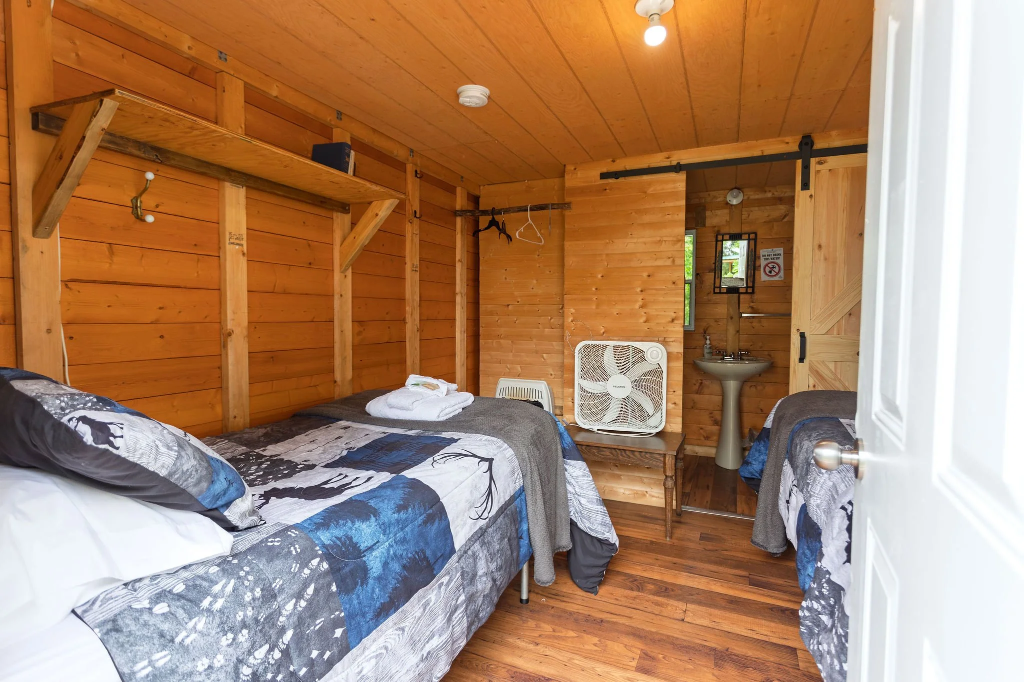 A rustic bedroom with wooden walls, a bed with patterned blue and gray bedding, a small wooden nightstand with a white fan, and a corner with a sink. There is a hanging rack with empty hangers and a shelf above with a few items. The room has a ceilin