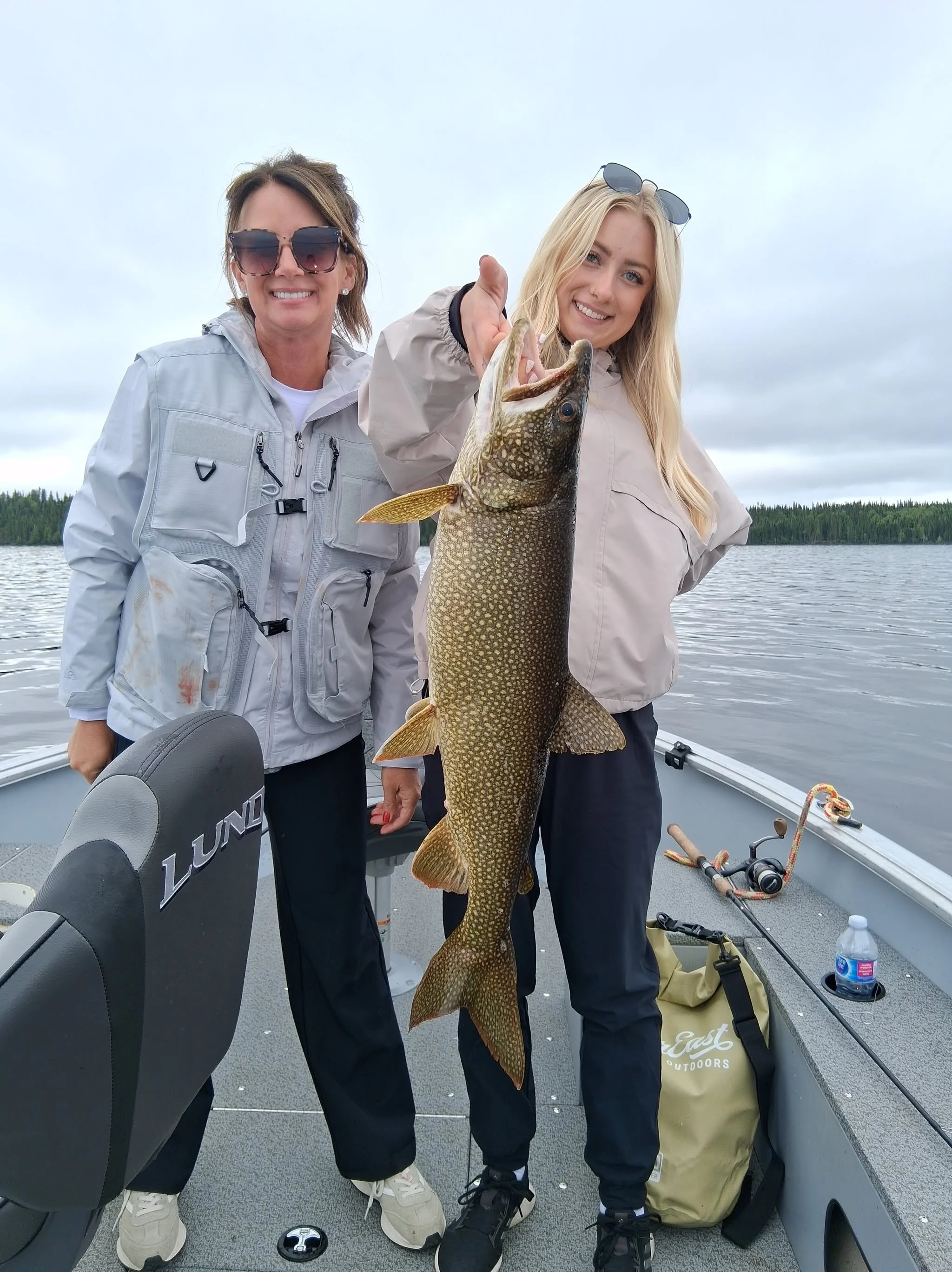 Two women on a boat holding a large fish, smiling. The woman on the left has short brown hair and sunglasses, wearing a gray jacket. The woman on the right has long blonde hair, is wearing sunglasses on her head, and a beige jacket. The background fe