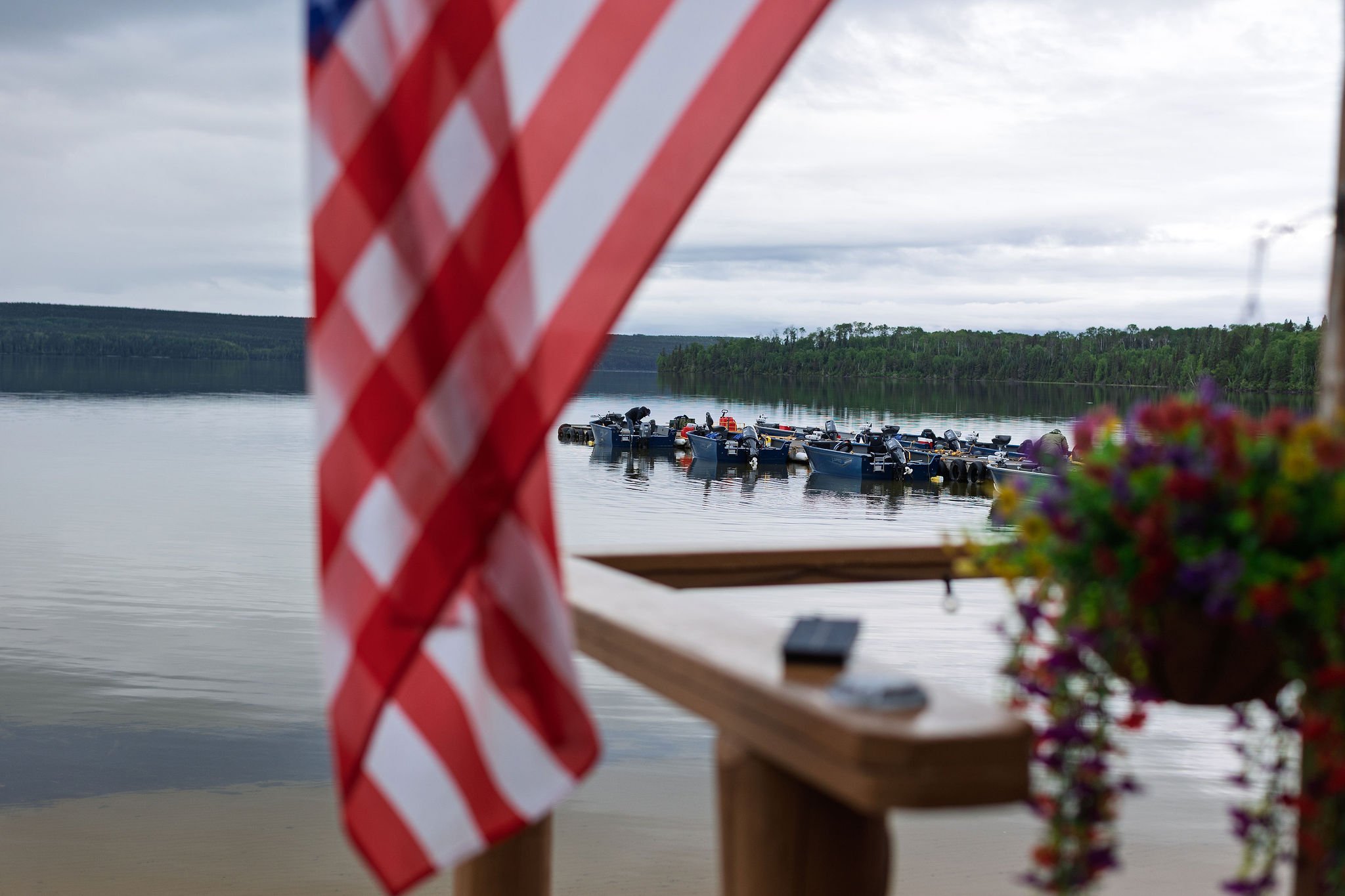 View of boats on a calm lake with overcast sky, as seen from a dock decorated with a red and white striped flag and potted flowers.