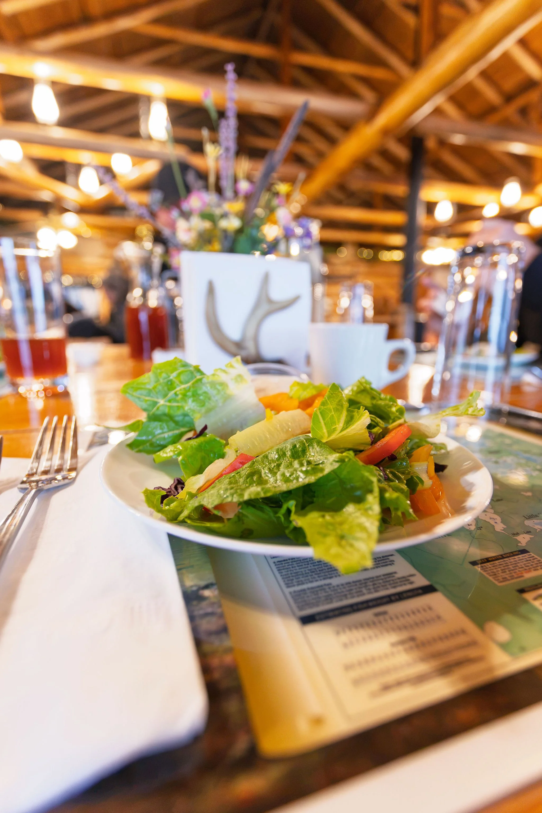 A plate of salad with leafy greens, tomatoes, and other vegetables on a table with a fork to the side. In the background, there are glasses, a cup, a napkin, and a floral arrangement in a vase, all set in a warmly lit restaurant with wooden beams.