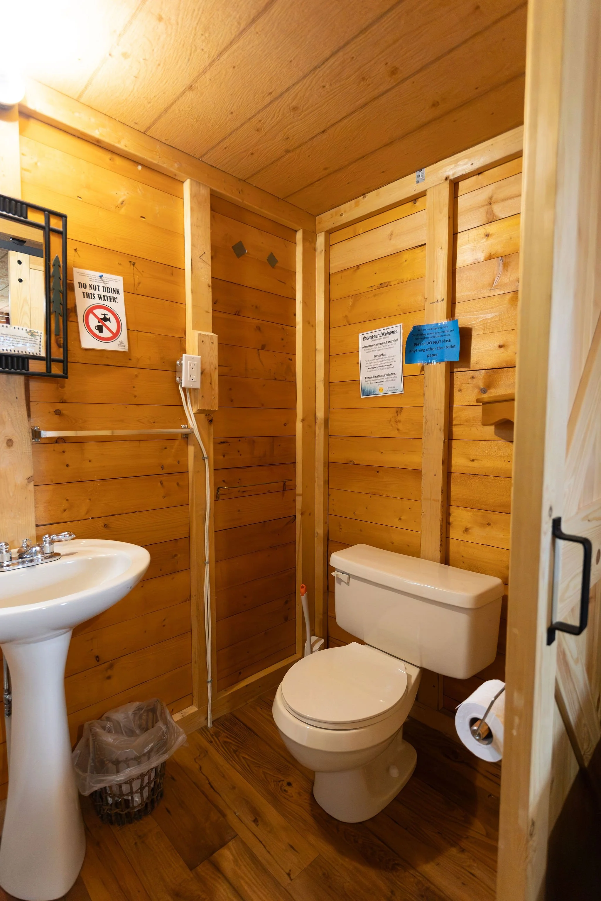 A wooden bathroom with a toilet, a pedestal sink, a trash can, and signs on the wall indicating not to drink the water and to not flush anything other than toilet paper.