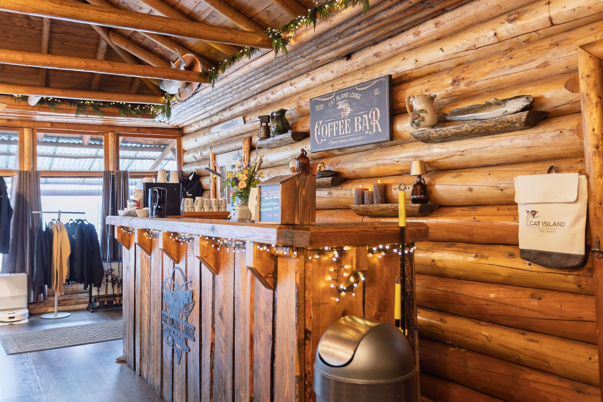 Interior of a rustic log cabin-style coffee shop with a wooden counter, decorative shelves holding vases and a fish figure, string lights, and a sign that reads 'Cat Island Lodge Coffee Bar'.
