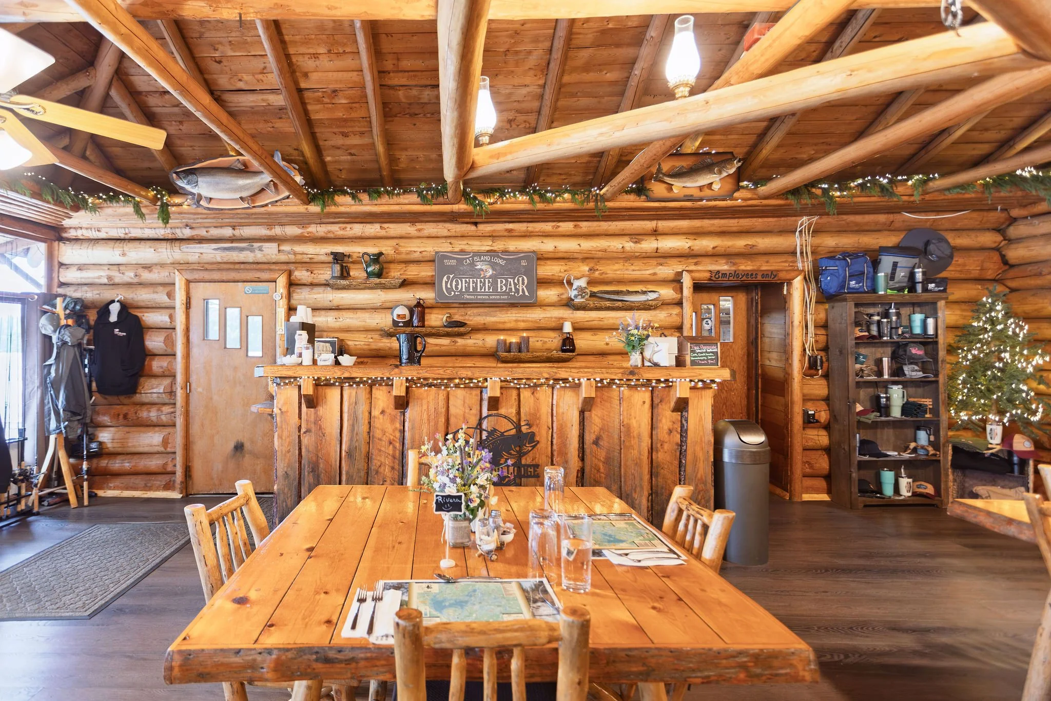 Interior of a rustic log cabin restaurant with wooden beams and walls, decorated with mounted fish, fairy lights, and greenery. Wooden tables and chairs are set for dining.