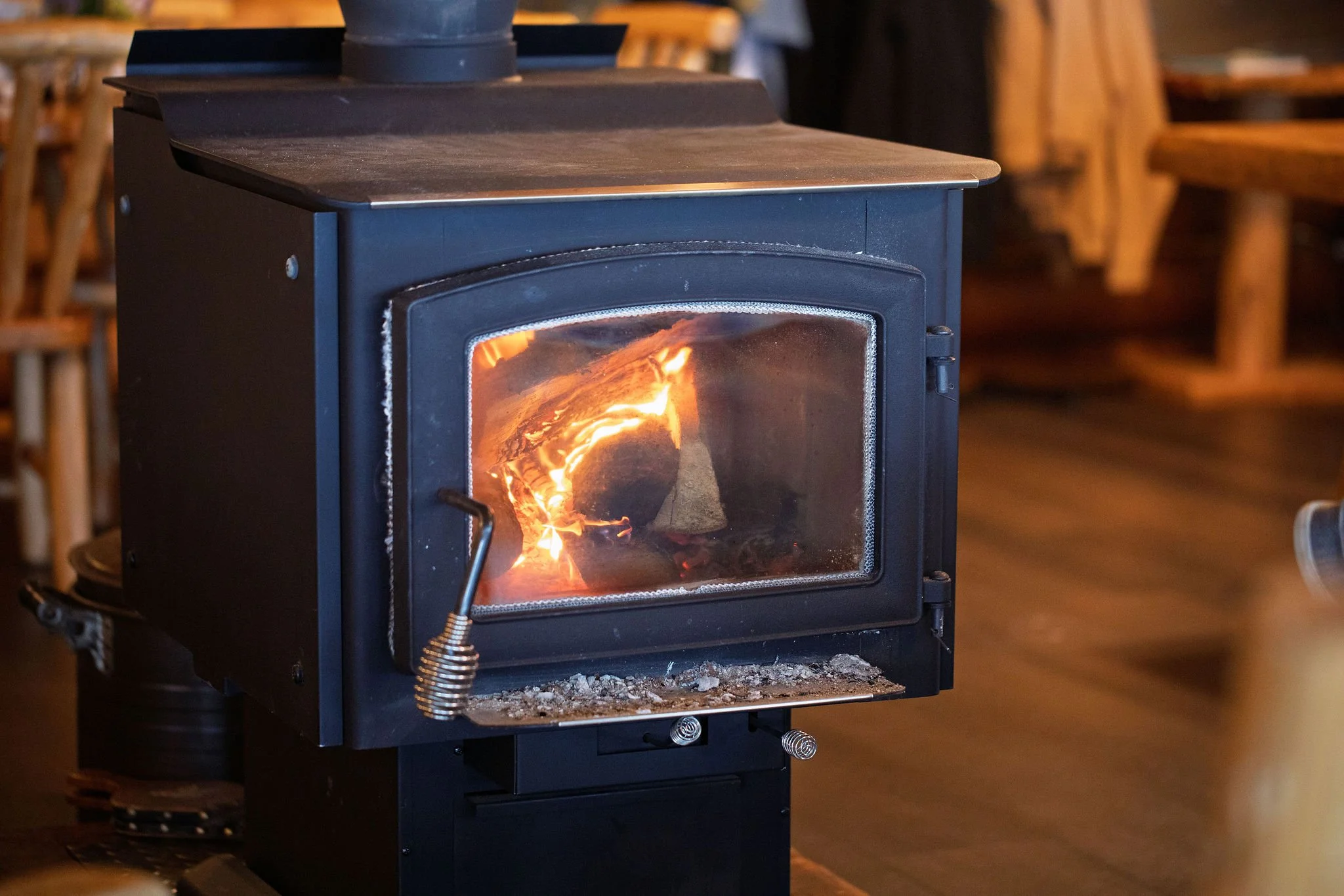 A black wood-burning stove with a glass door showing a visible fire inside, situated in a cozy indoor setting with wooden furniture in the background.