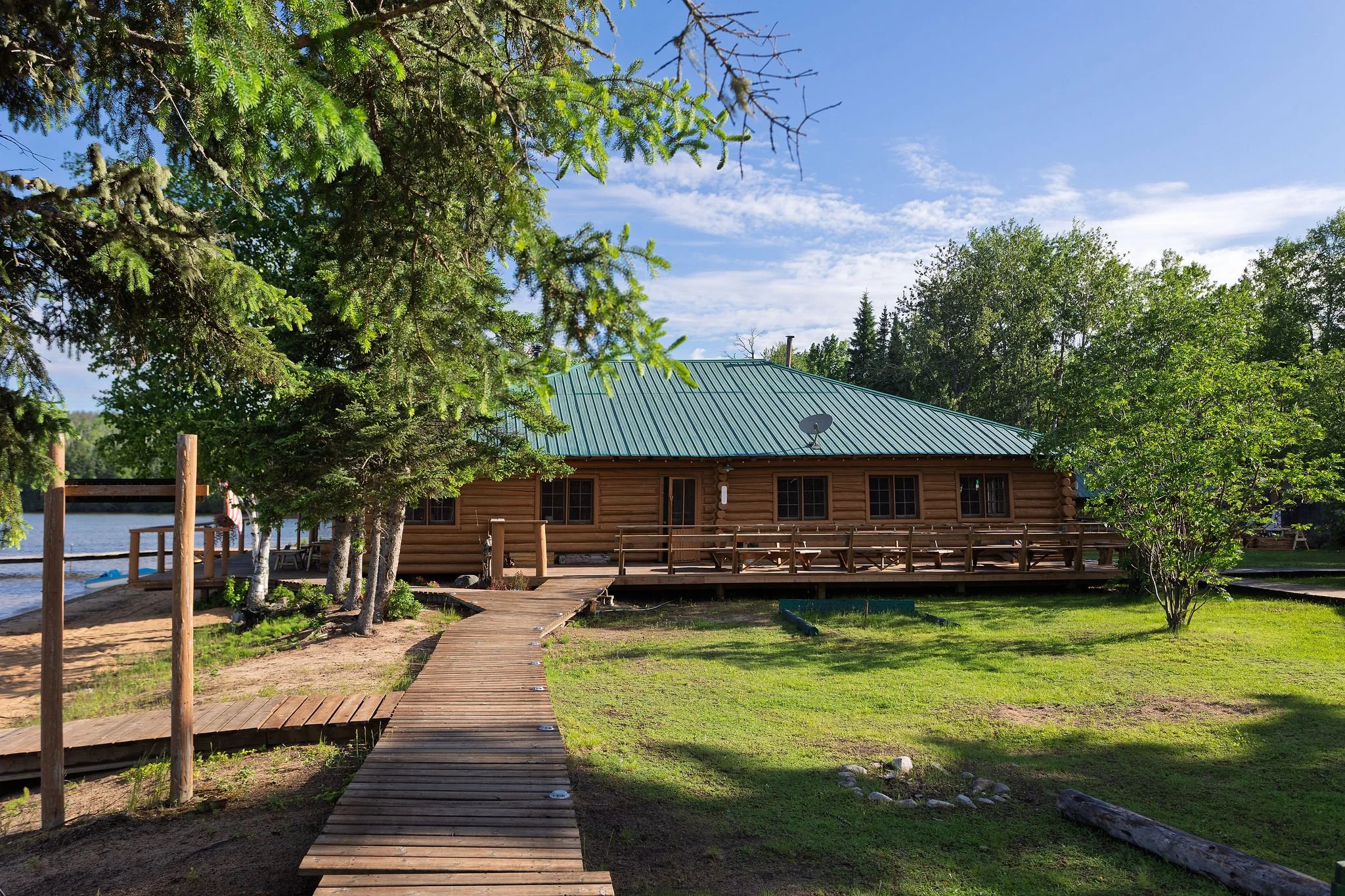 A wooden cabin with a metal roof is situated near a body of water, surrounded by green trees. A wooden walkway leads to the cabin, with a satellite dish on the roof. The sky is partly cloudy with a clear blue tone.