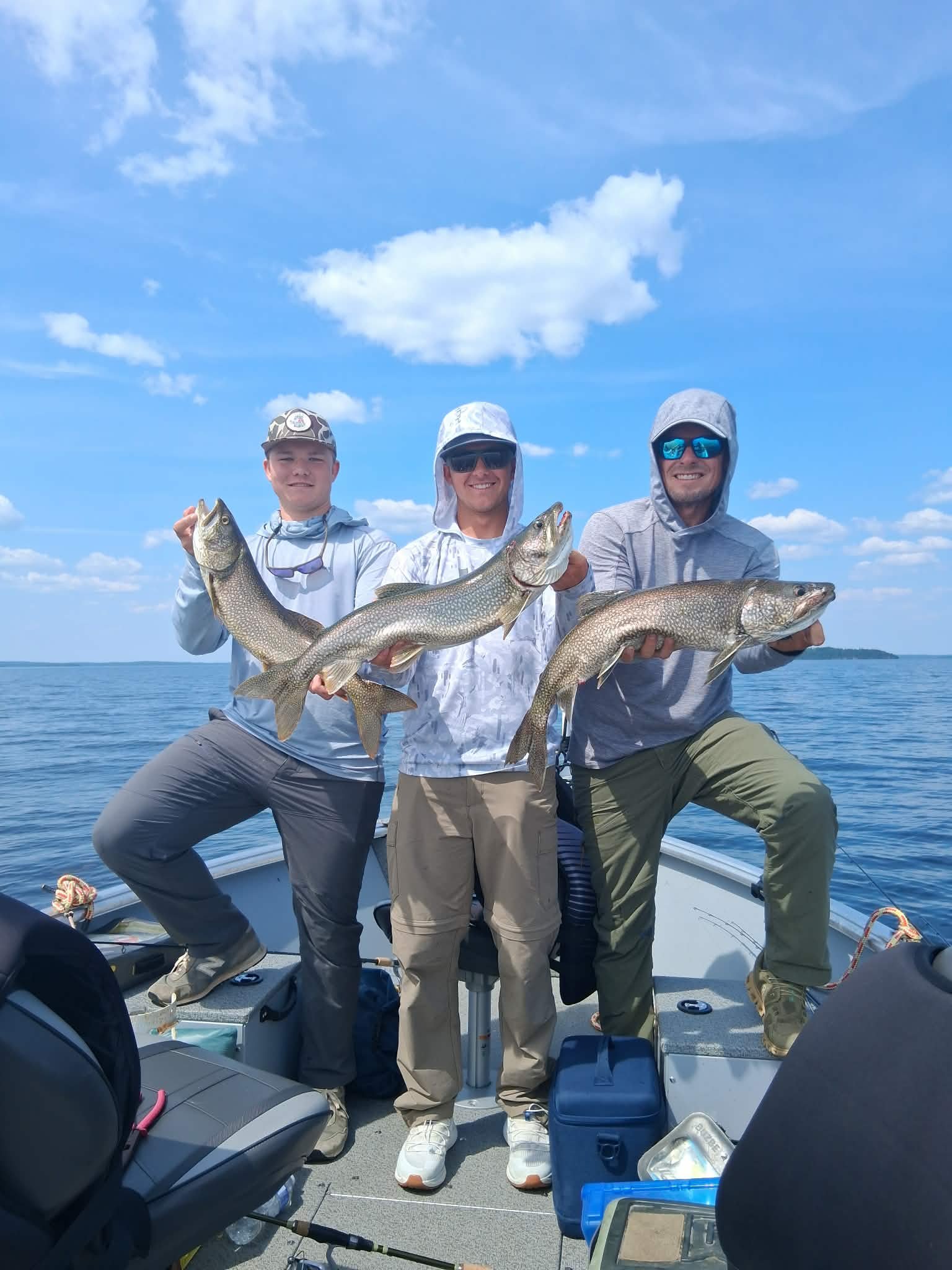 Three men in boat holding large fish, smiling, with blue sky and clouds in background.