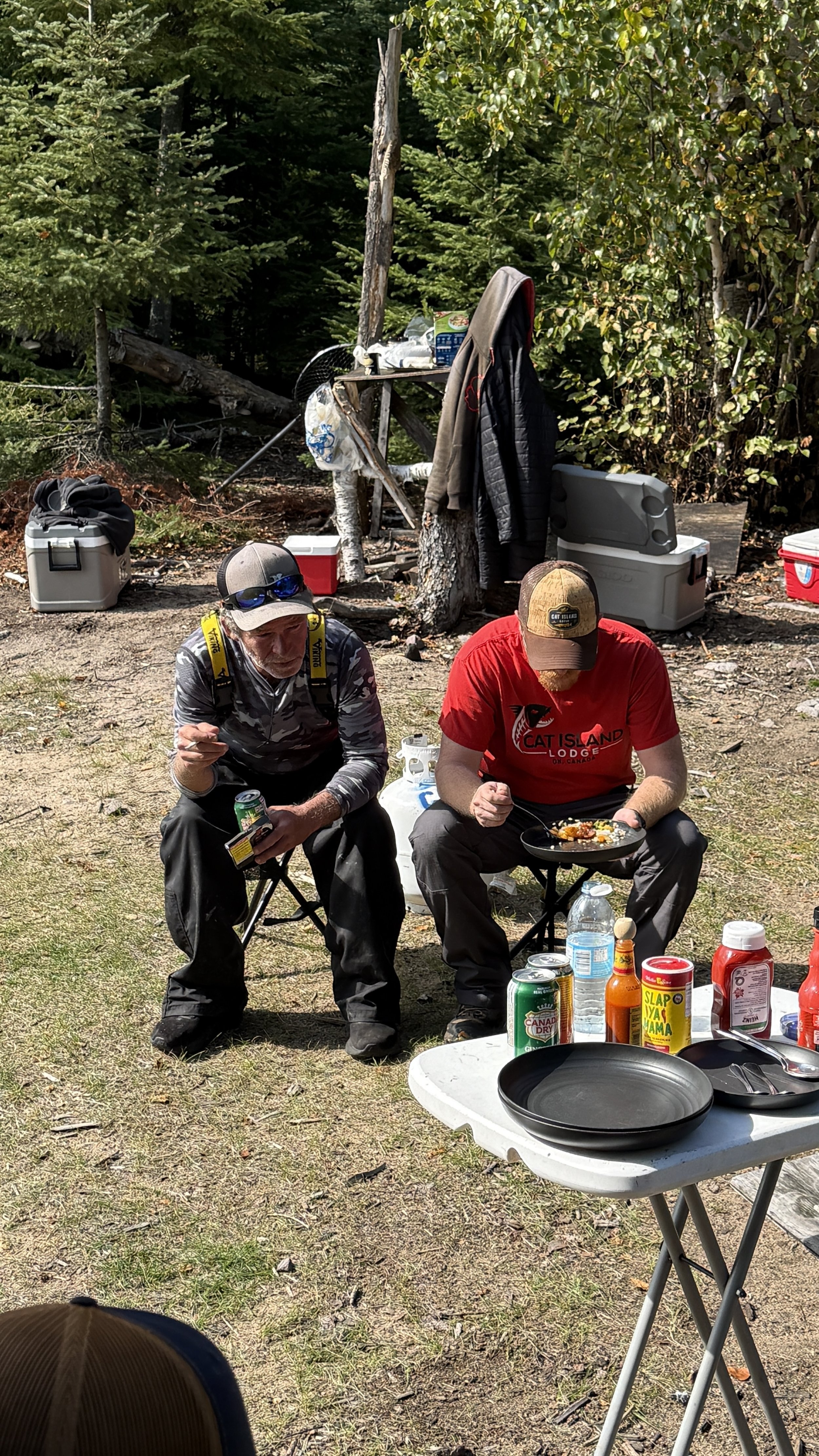 Two men sitting outdoors at a shore lunch on cat island in ontario, eating fresh caught fish with camping supplies and coolers in the background, surrounded by trees.