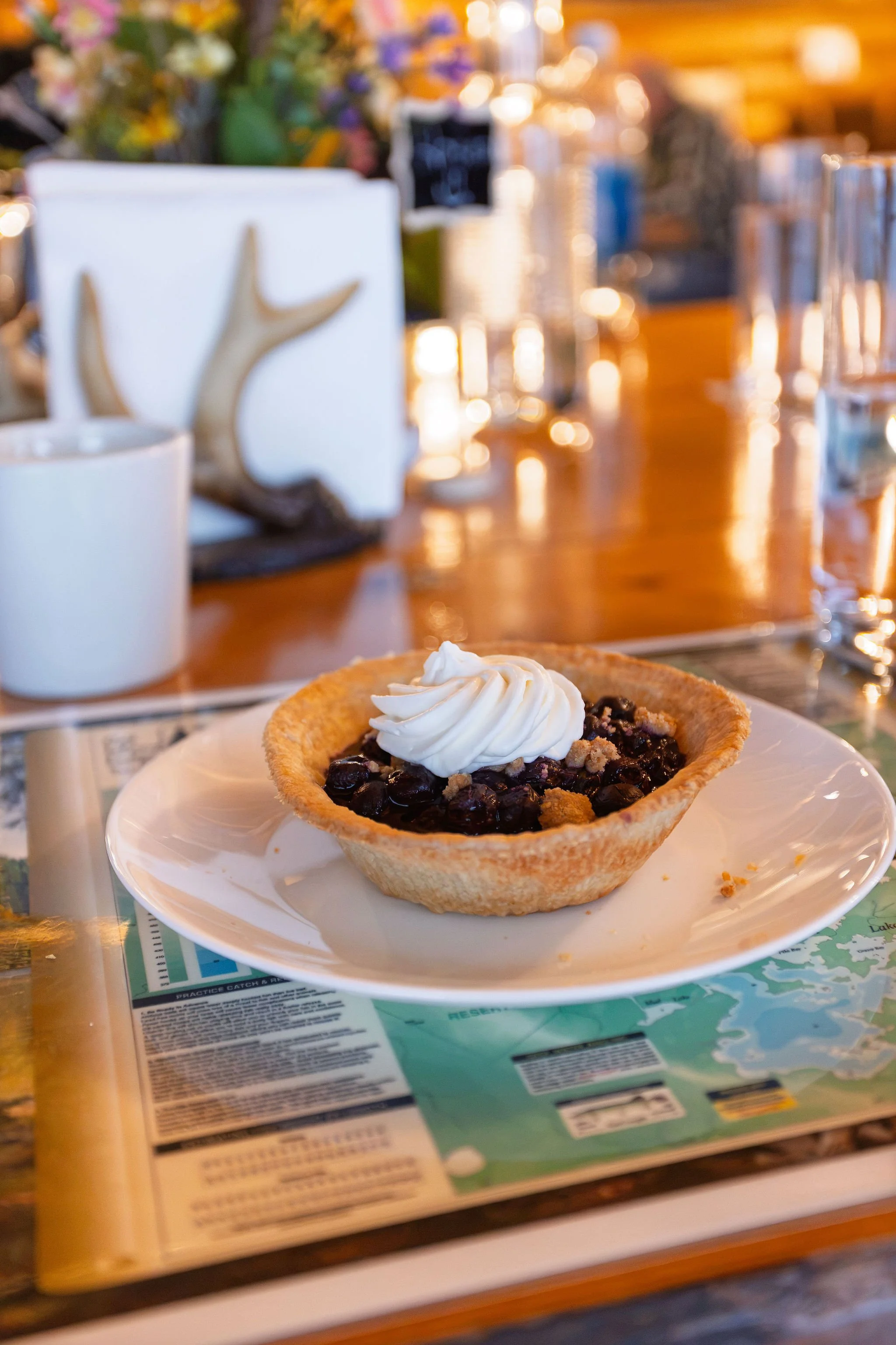 A blueberry tart garnished with whipped cream on a white plate, placed on a table with a menu underneath. The background features a blurred restaurant setting with colorful flowers and glasses.