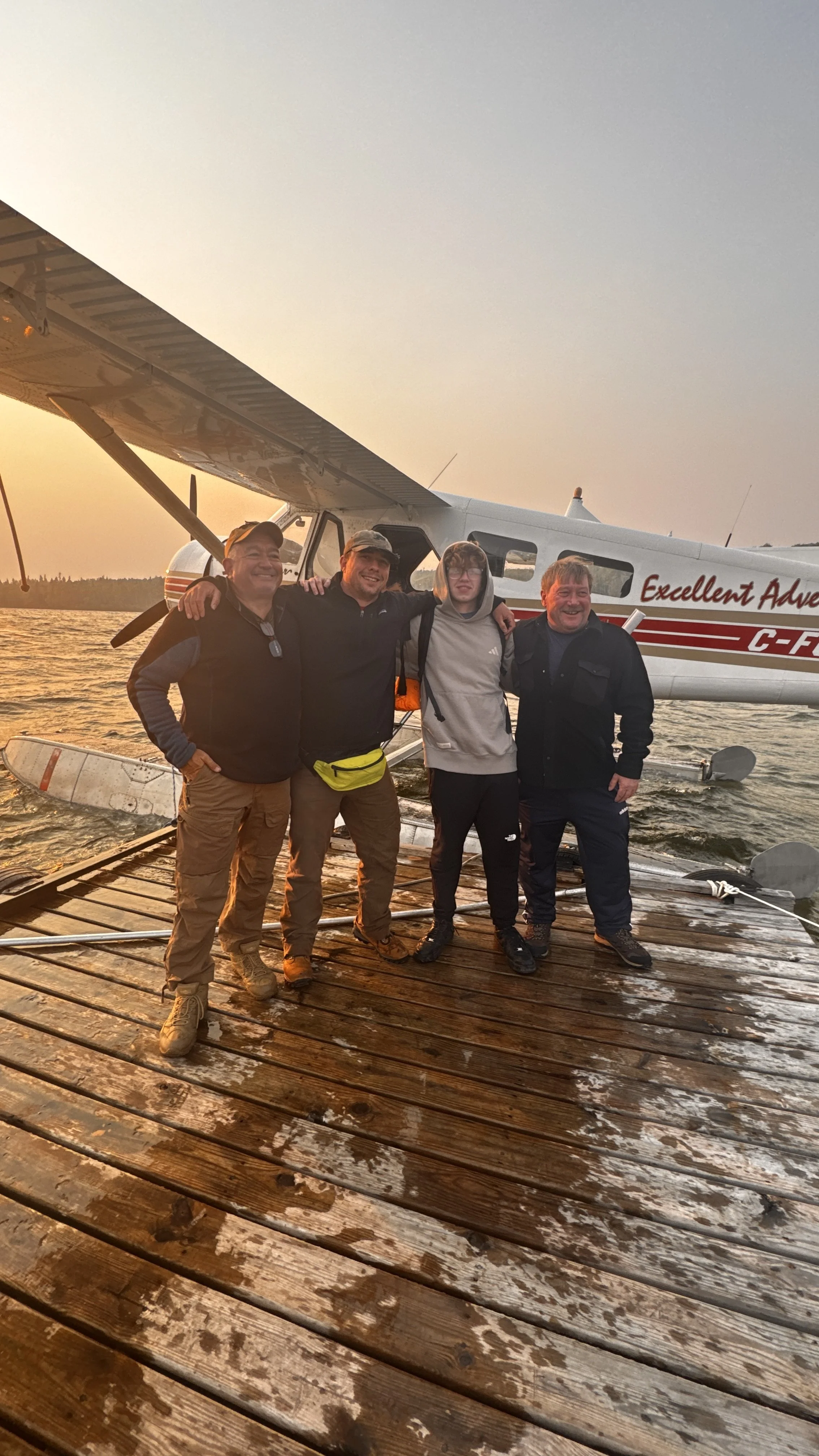 Four men standing on a wooden dock on trout lake in front of a small seaplane during sunset, smiling and with arms around each other.