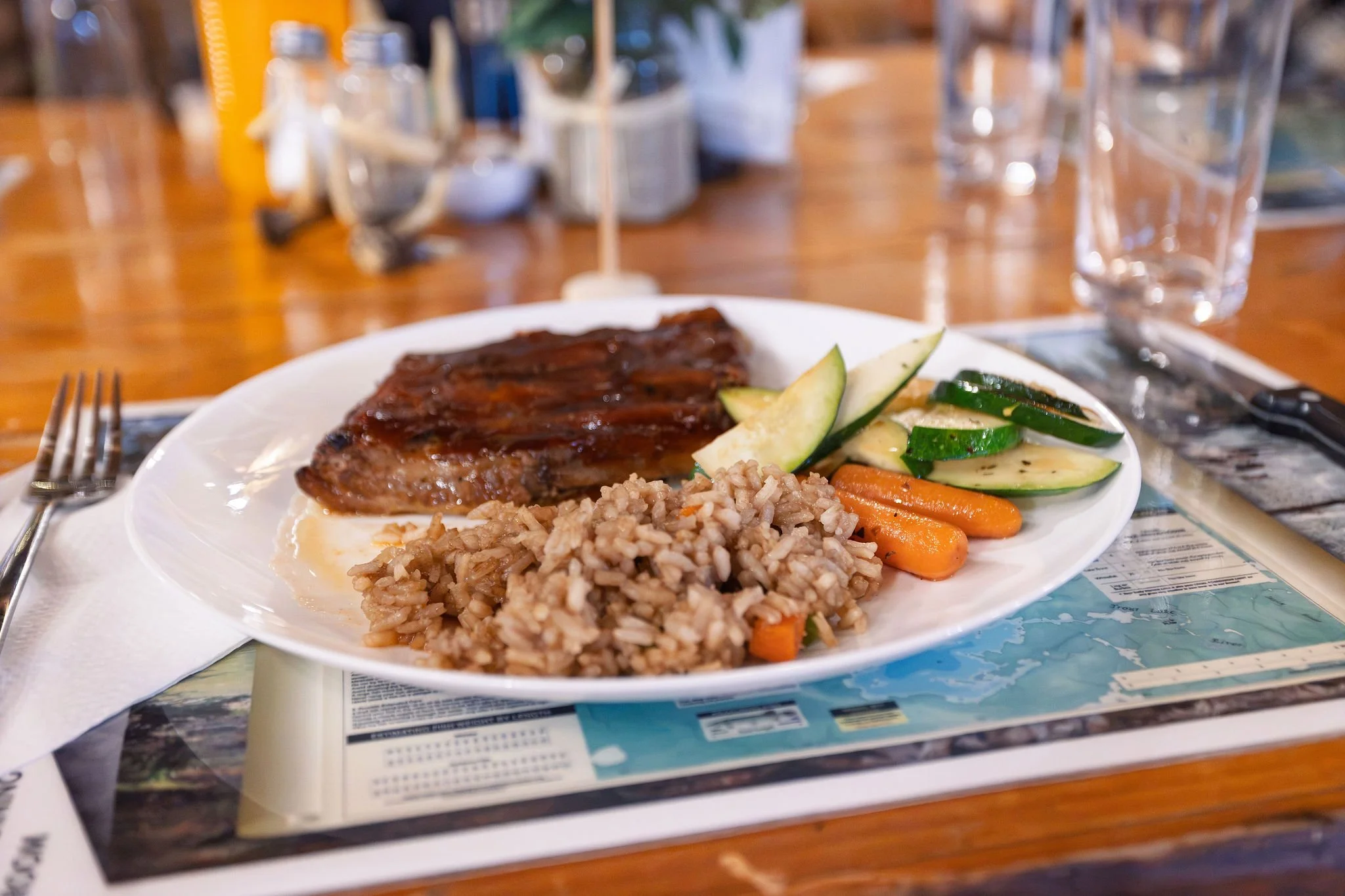 A white plate with baked chicken, cooked rice, and a mix of sliced cucumbers and carrots. The plate is on a table with a fork on the left and a knife and glass of water on the right, with a blurred background of a wooden dining table and various cond