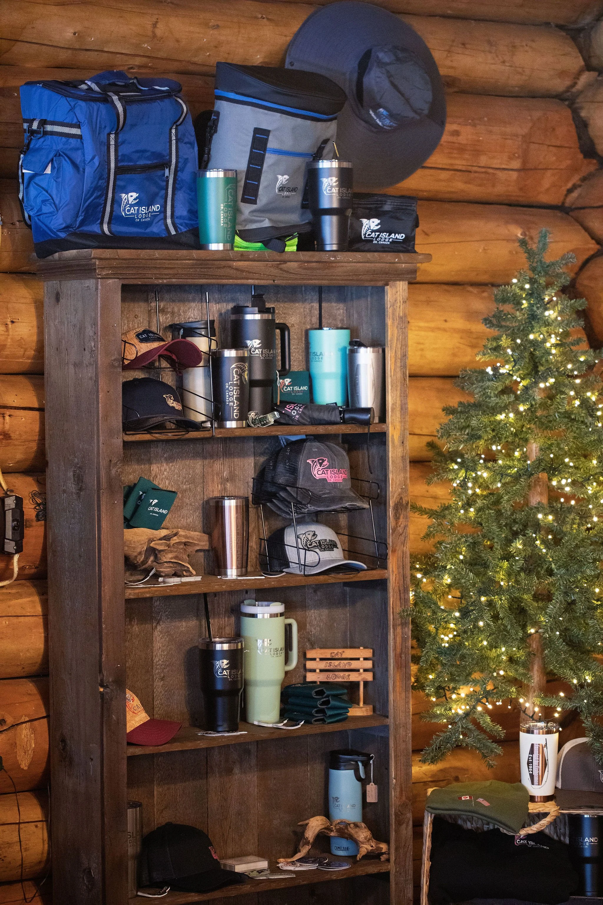 Wooden bookshelf with camping gear and accessories, including hats, mugs, thermoses, and a small artificial Christmas tree with lights.