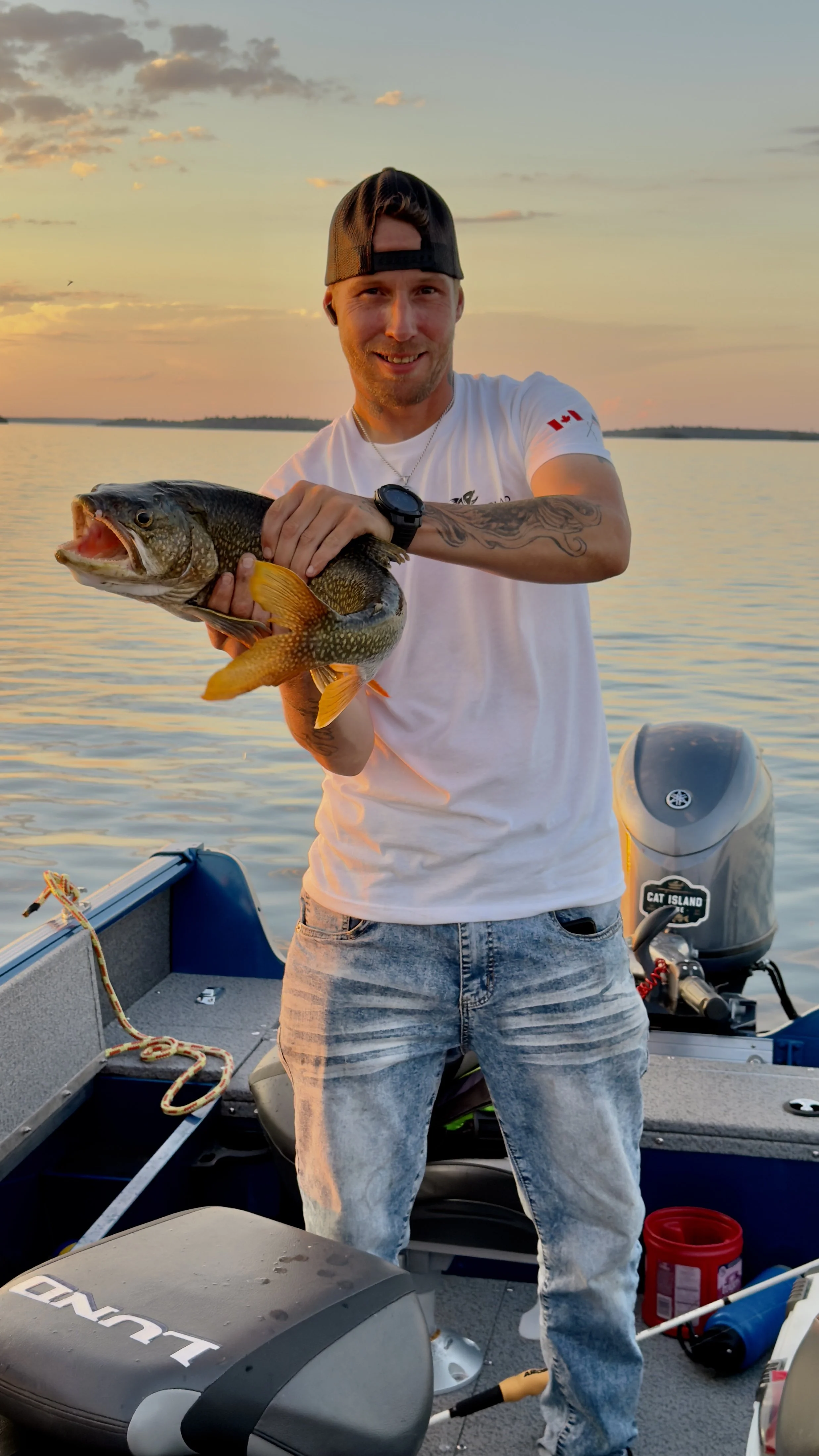 A man in a white T-shirt and jeans holding a large fish on a boat during sunset.