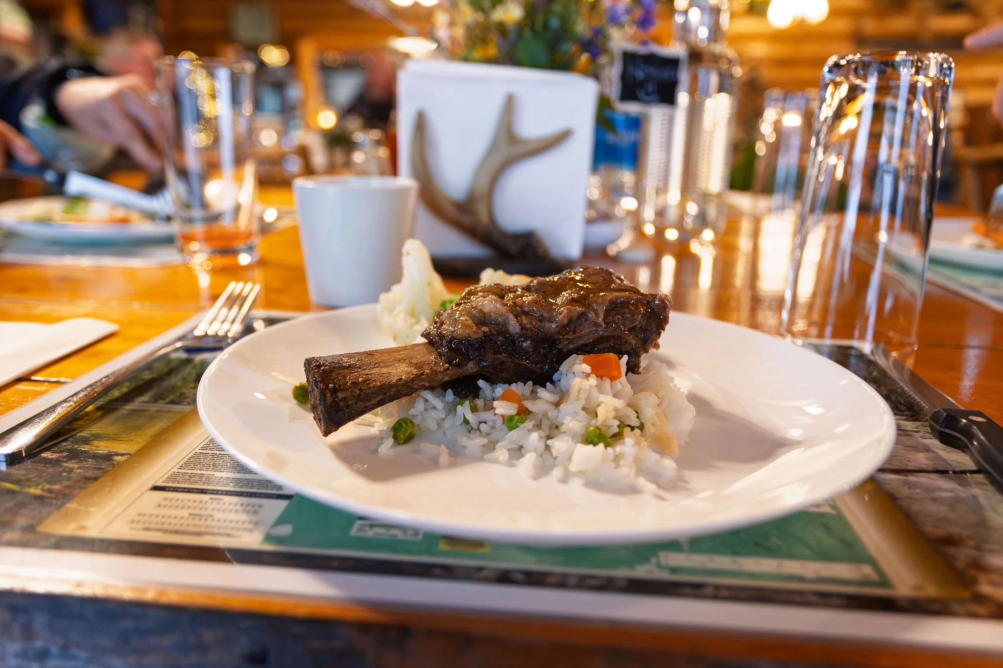A plate of food with rice, a beef short rib, and mashed potatoes on a table set indoors with various drinking glasses, utensils, and decorative items in the background.