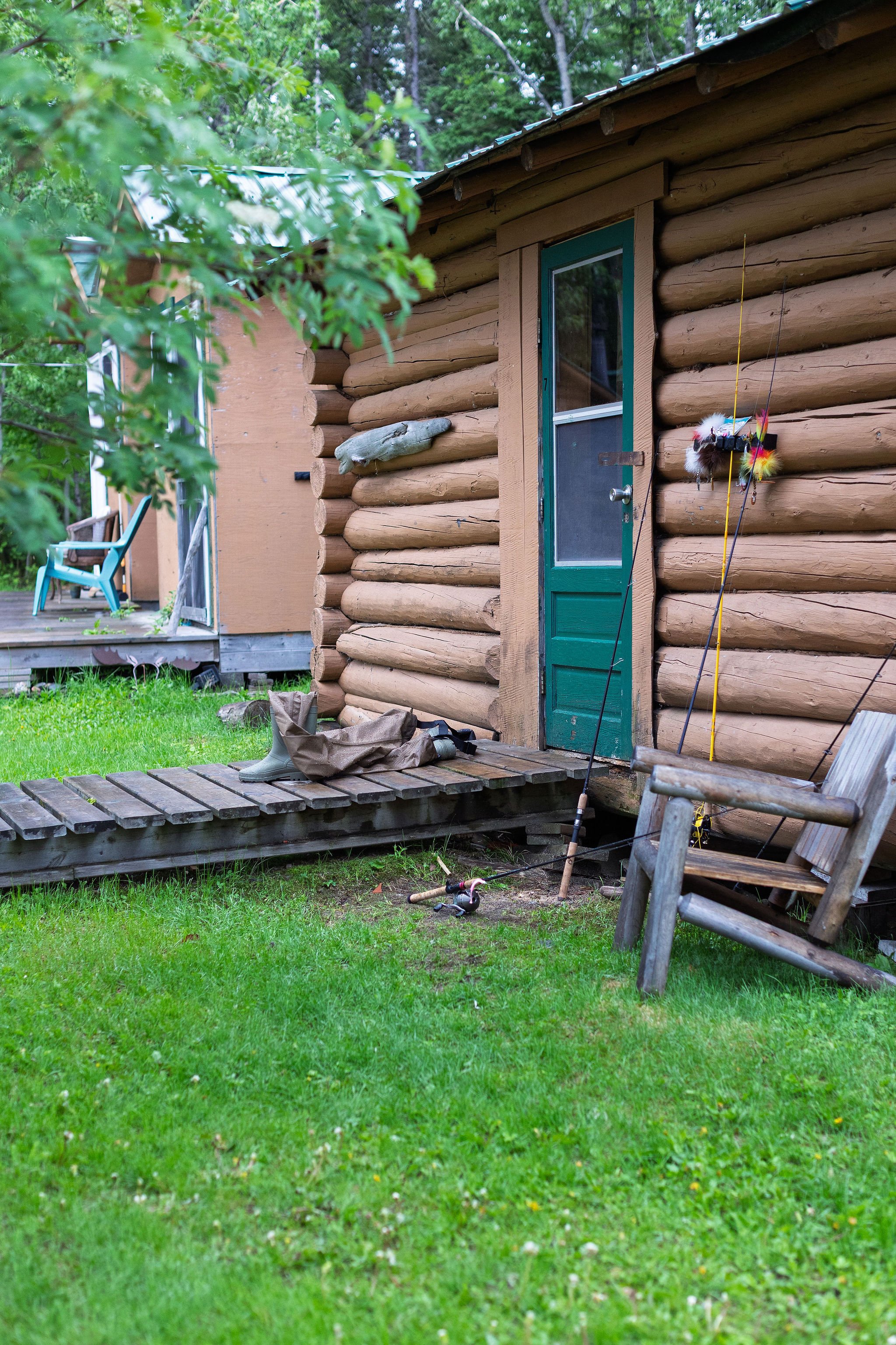 A rustic log cabin with a green door in a wooded area. The cabin has a small wooden porch with a rain boot, a bag, and some tools on it. There is a chair near the cabin and fishing poles leaning against the wall. Lush green grass and trees surround t