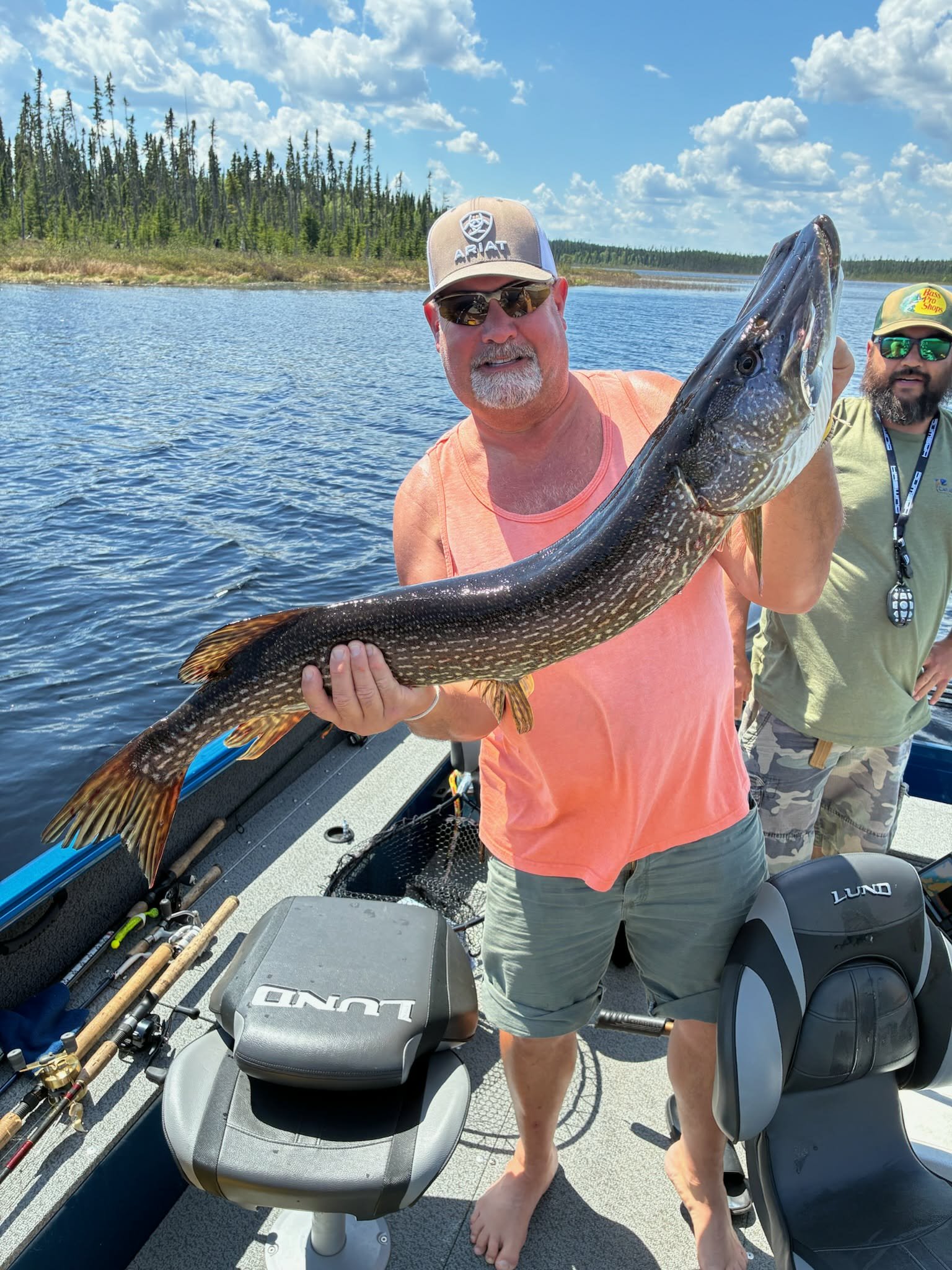 A man holding a large fish on a boat with fishing equipment and two other men in the background, on a lake with a forested shoreline under a partly cloudy sky.