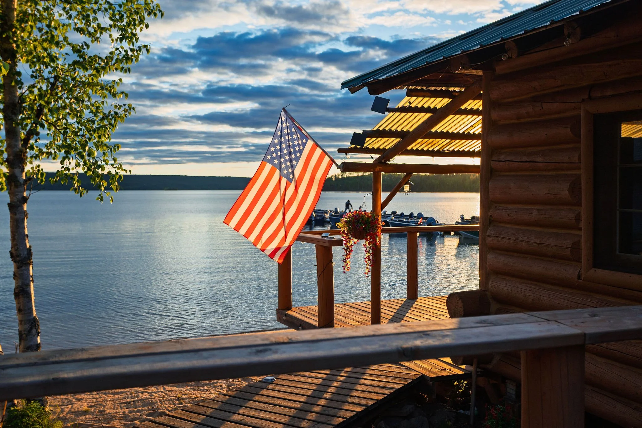 A wooden cabin balcony overlooking a lake with an American flag hanging from the railing. Trees are visible on the left, and boats are moored in the water. The sky is partly cloudy during sunset or sunrise.