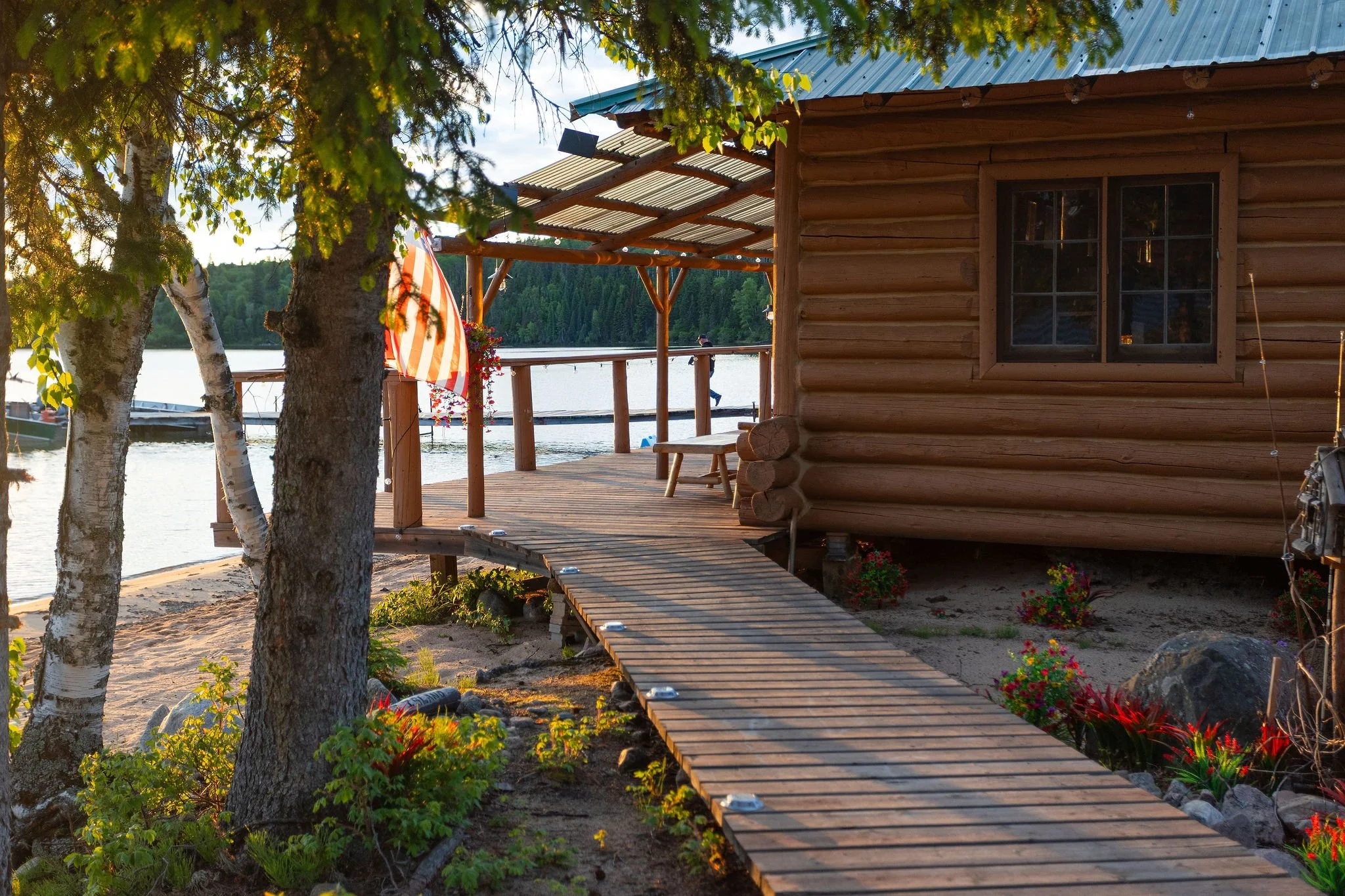 Cat Island Lodge on Trout Lake, Ontario, with a wooden deck, trees in the foreground, and a person fishing in the background. There are colorful flowers at the base of the deck and an American flag next to the cabin.