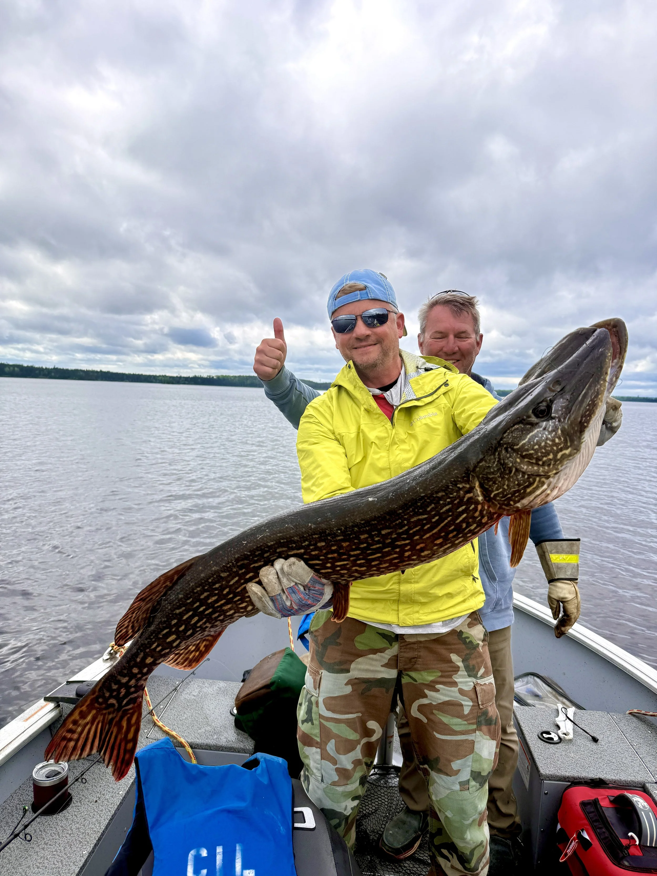 Two men on a boat holding a large fish with dark markings, under cloudy skies, with water and a distant shoreline in the background.