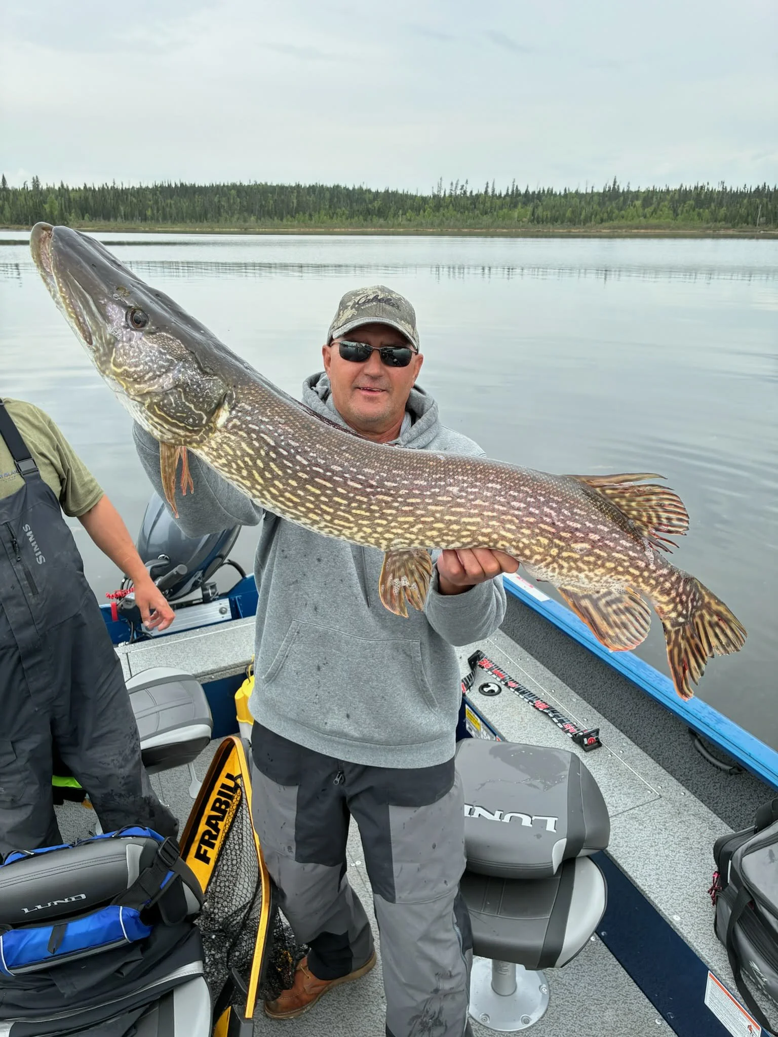A man wearing sunglasses, a cap, and gray hoodie holding a large fish, possibly a northern pike, on a boat on a calm lake with a forest in the background.