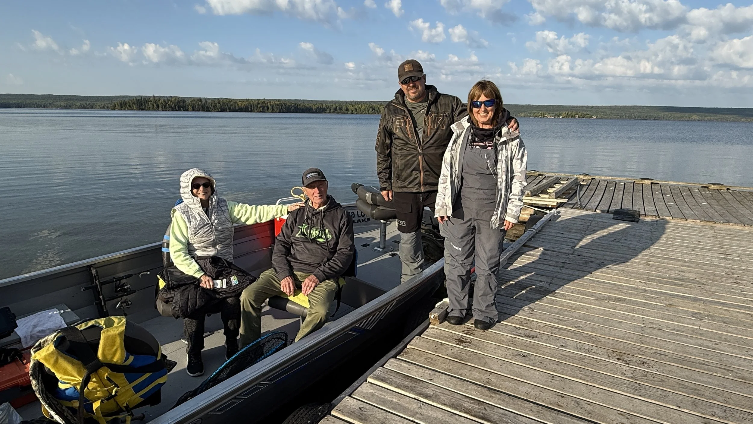 Four people on a boat and a dock by a lake at Cat island lodge, smiling for the photo. The group includes two women and two men, all dressed in outdoor gear with sunglasses, with a shoreline and blue sky in the background.