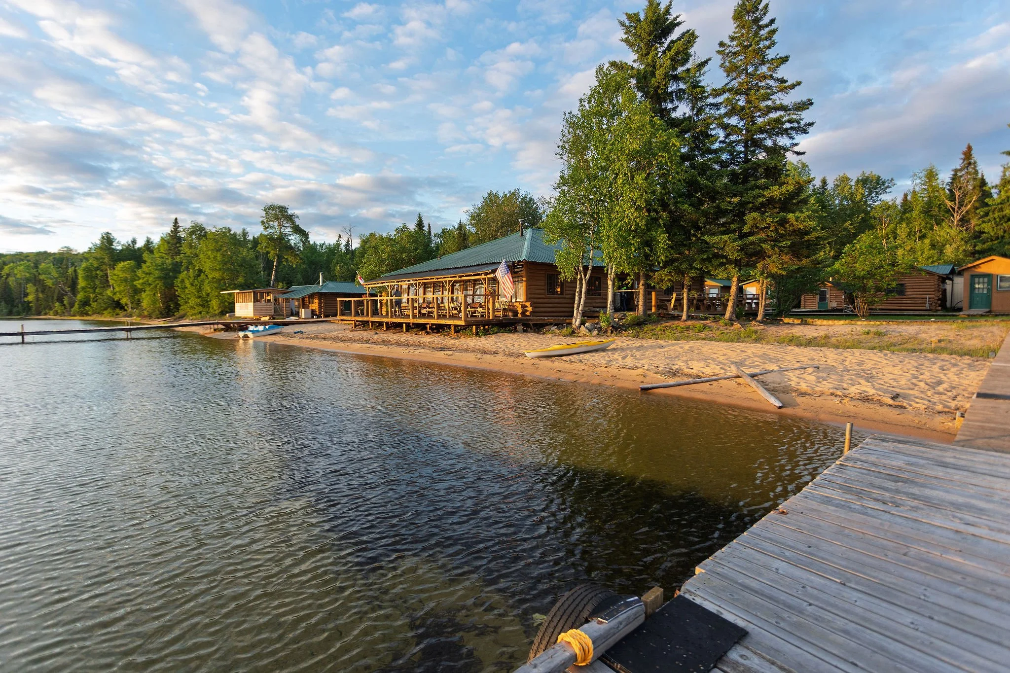 A lakeside scene with a wooden dock extending into the water, bordered by trees and small cabins in the background. The sky is partly cloudy with the sunlight casting a warm glow on the landscape.