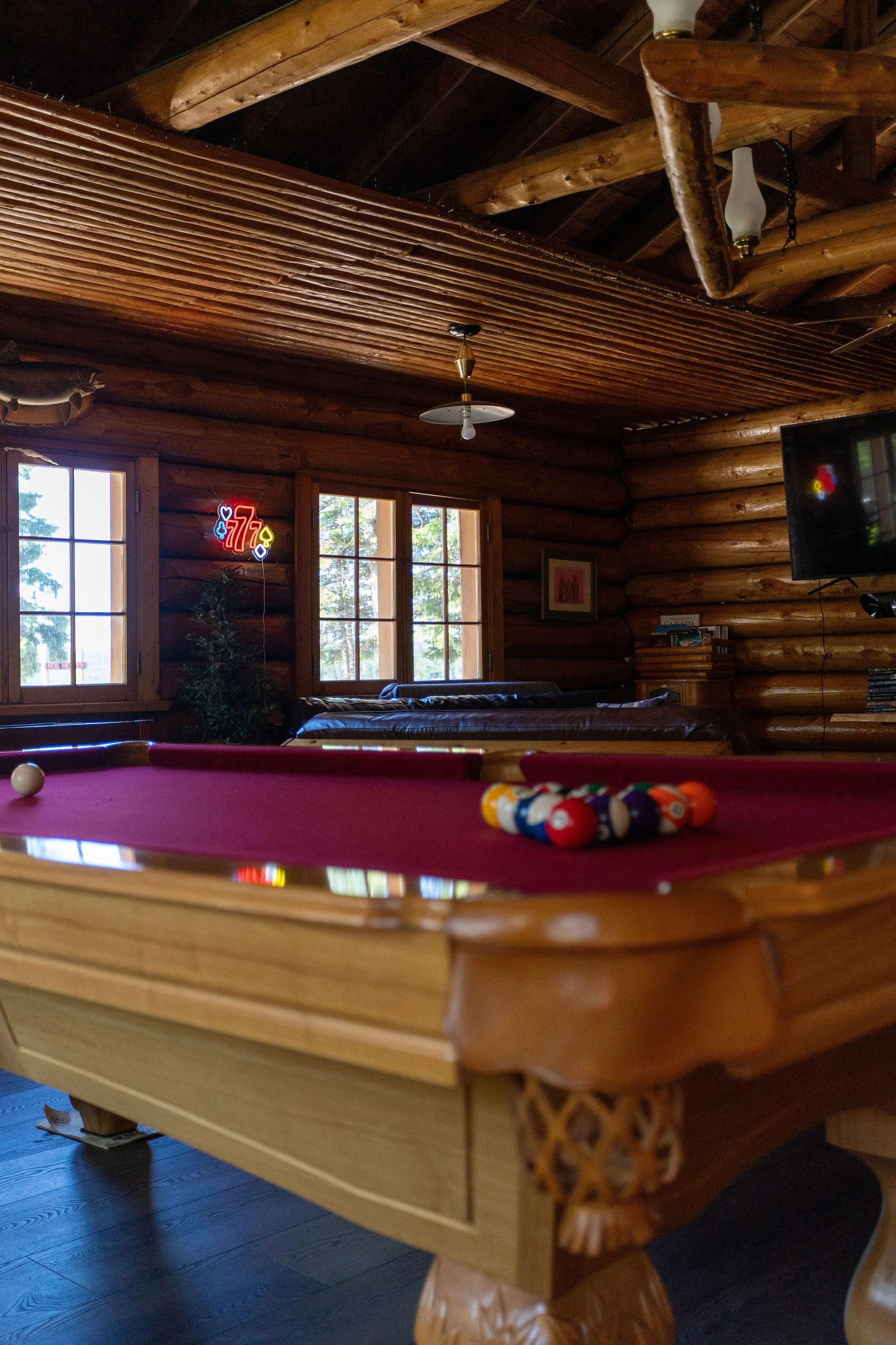 Interior of a log cabin with a pool table in the foreground, windows letting in natural light, wooden beams on the ceiling, a TV on the wall, and a neon sign.