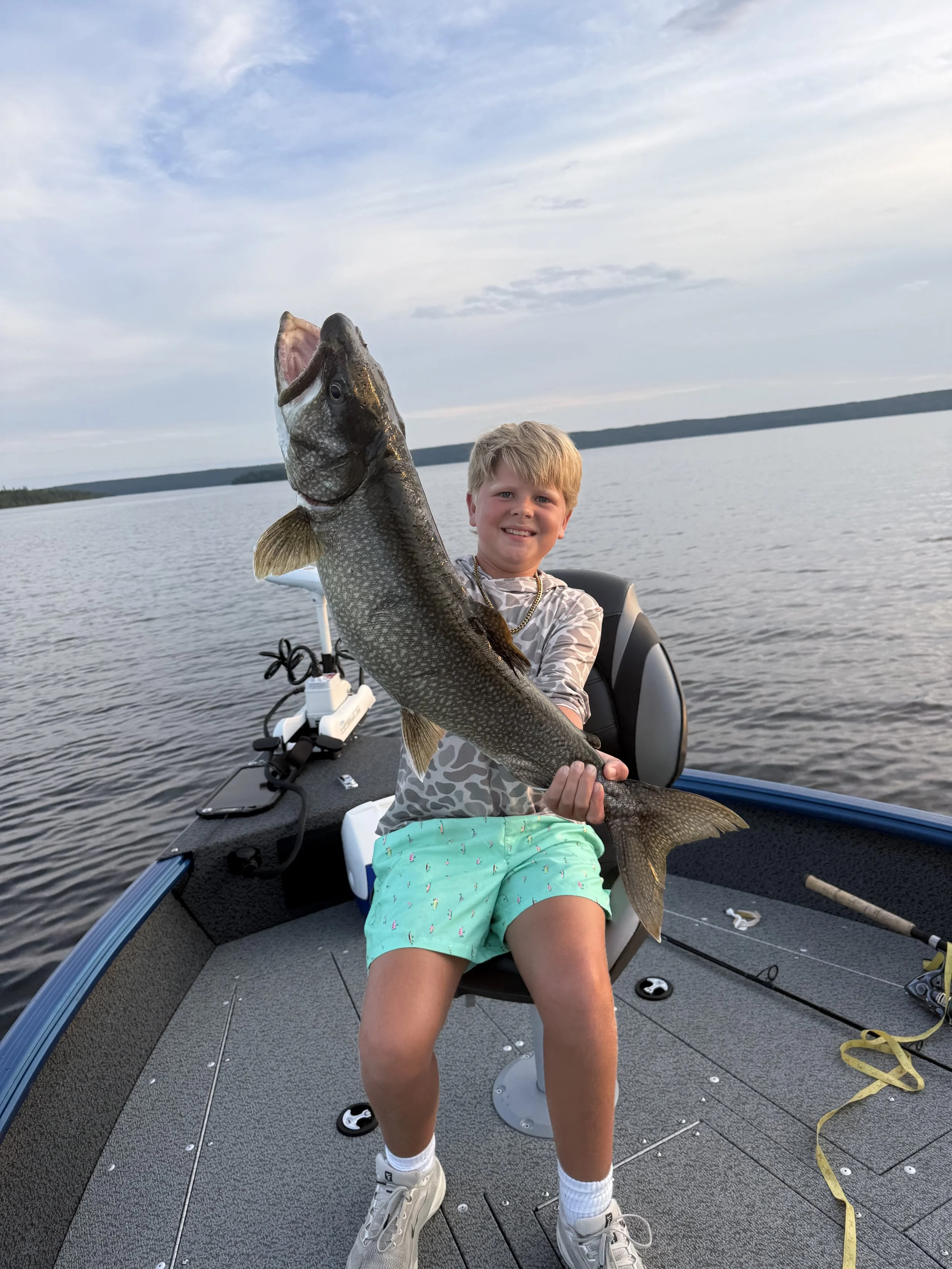 A young boy on a boat holding a large fish he caught, with water and distant shoreline in the background.