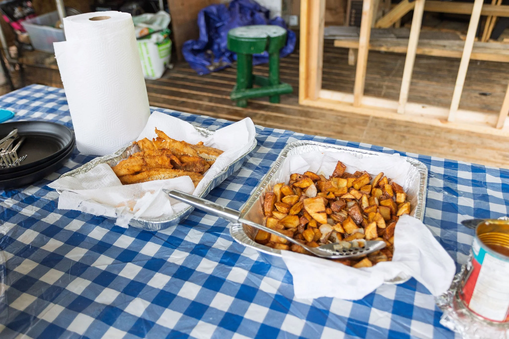 Fried fish fillets and cooked diced potatoes served in aluminum trays on a table with a blue and white checkered tablecloth, in a partly built wooden structure.
