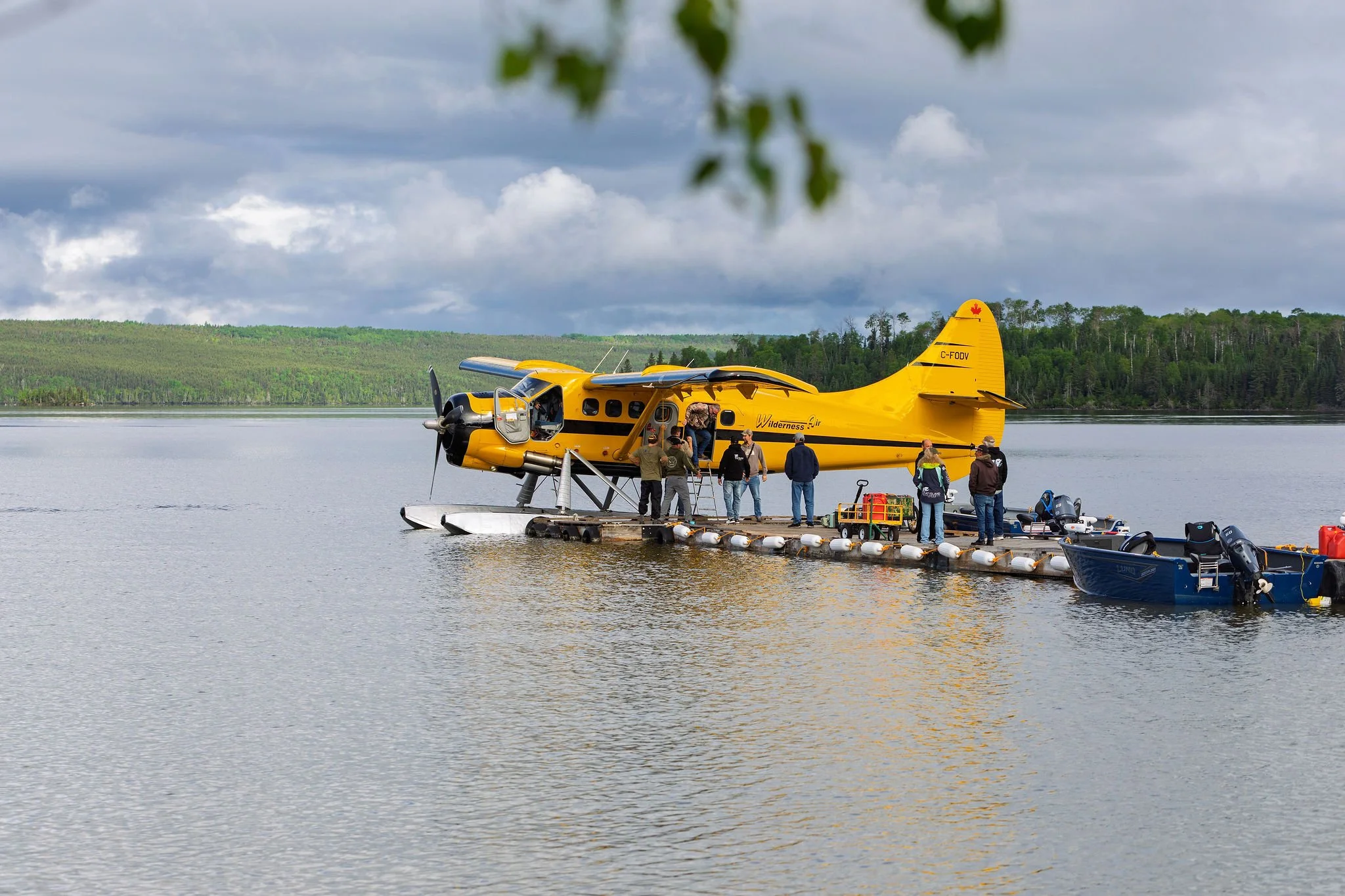 A bright yellow seaplane docked at a floating platform on a lake, with people boarding and equipment nearby, surrounded by green forest and cloudy sky.