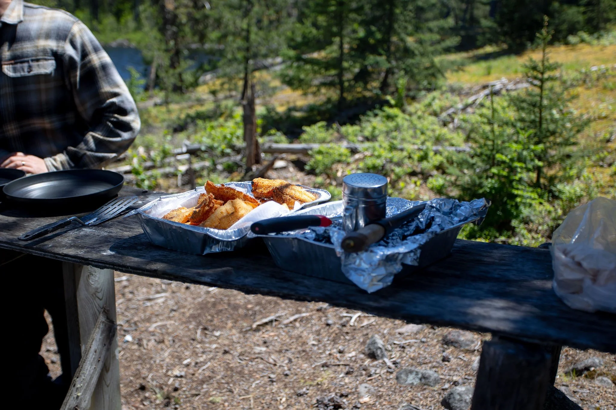Fried fish fillets on a foil-lined tray sitting on a rustic outdoor wooden table in a forested area.