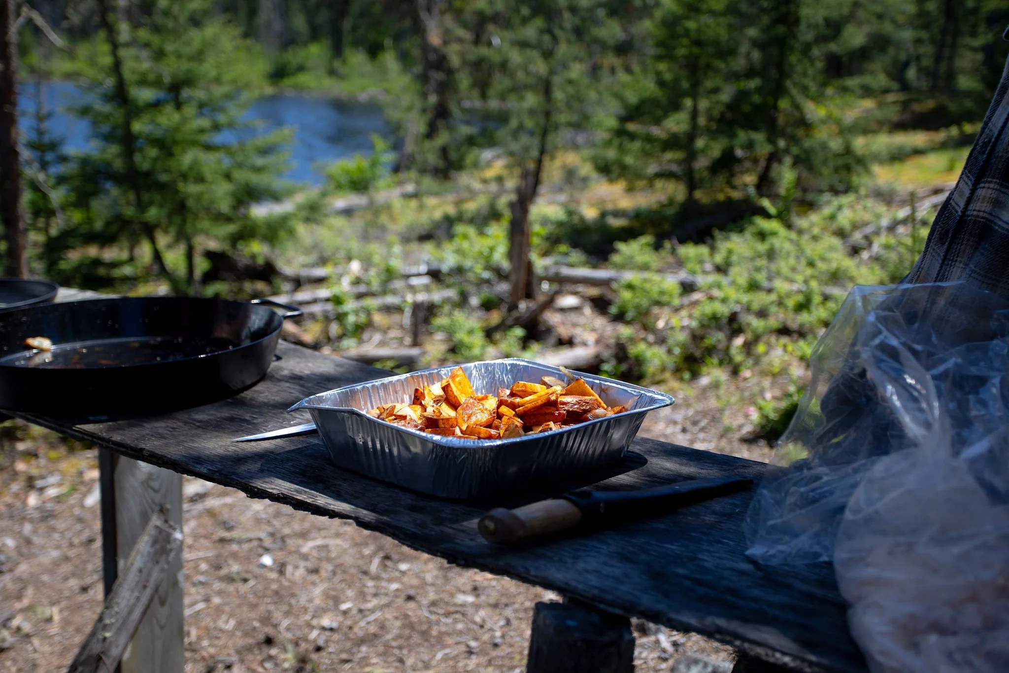 Outdoor scene with a table set up in a forest. On the table, there is a metal tray of cooked sweet potatoes and a black cast-iron skillet. The background shows trees, a lake, and greenery.