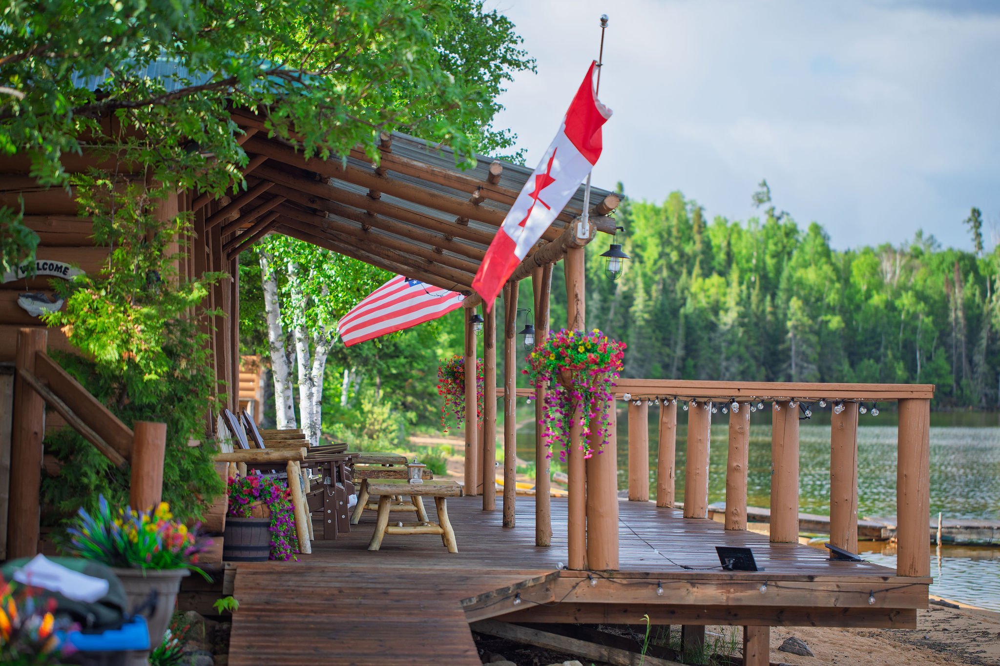 The wooden lakeside deck of Cat Island Lodge in Ontario decorated with Canadian and American flags, hanging flower baskets, string lights, and surrounded by lush green trees.