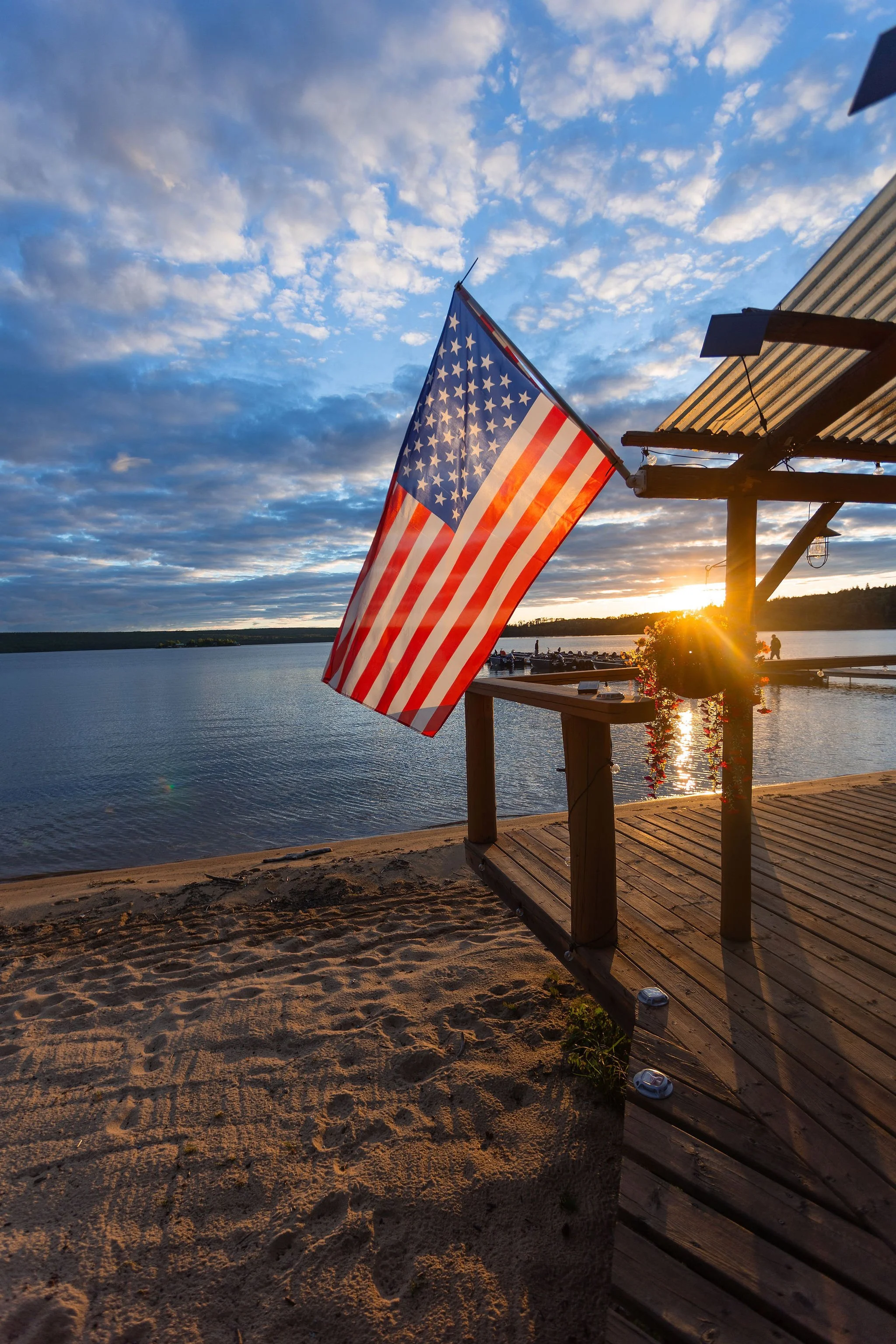 American flag hanging on the wooden deck of Cat Island Lodge overlooking Trout Lake during sunset with a cloudy sky.