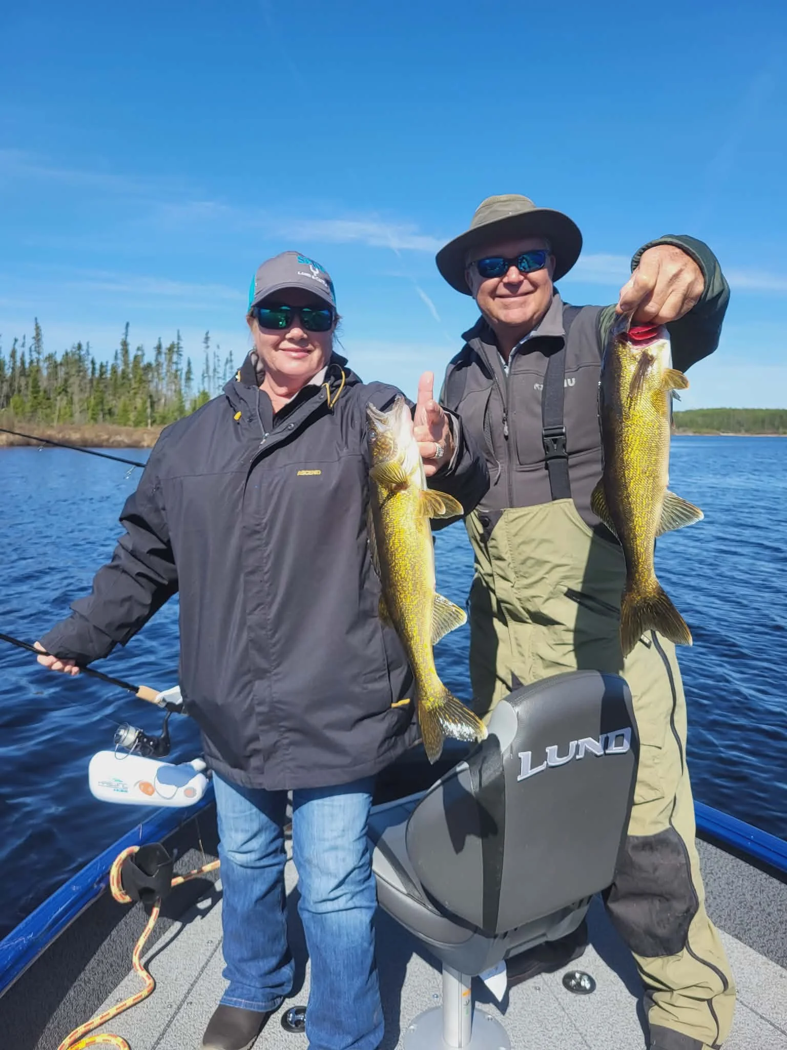 Two people on a boat, smiling and holding up large fish they caught during a fishing trip on a sunny day with blue sky and water in the background.